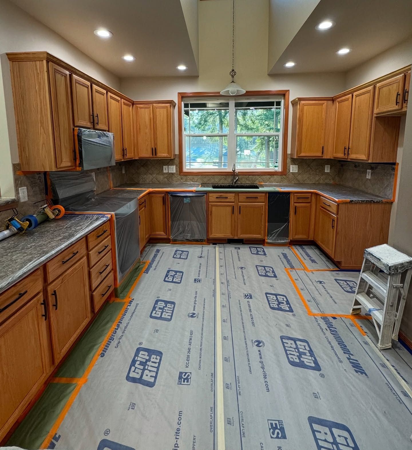 A U-shaped kitchen with natural oak cabinets, granite countertops, and protective flooring paper during a renovation.
