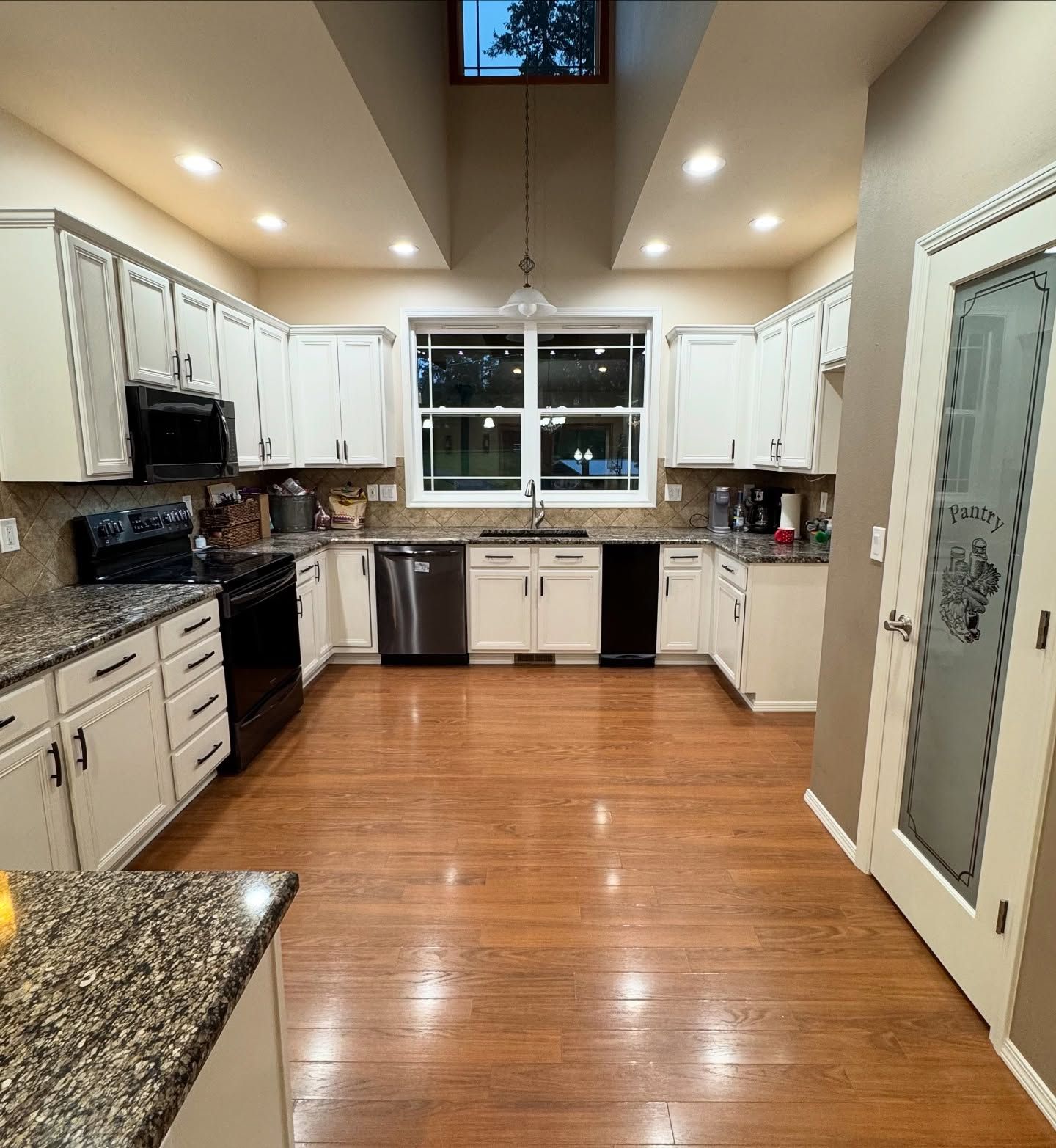 A well-lit kitchen with white cabinets, granite countertops, wood floors, and a pantry with a frosted glass door.