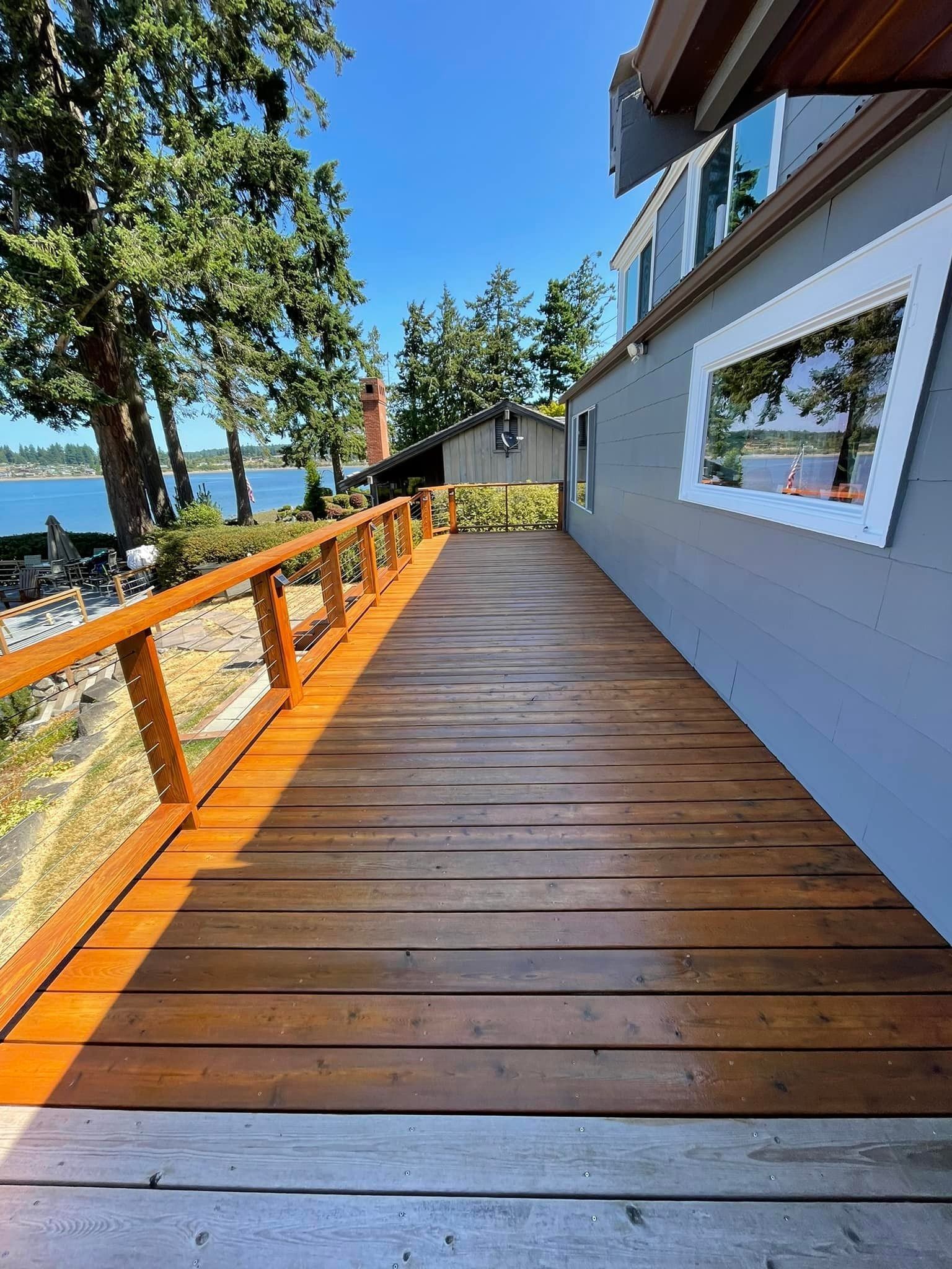 A long wooden deck runs along the side of a grey house, overlooking a body of water with trees in the background.