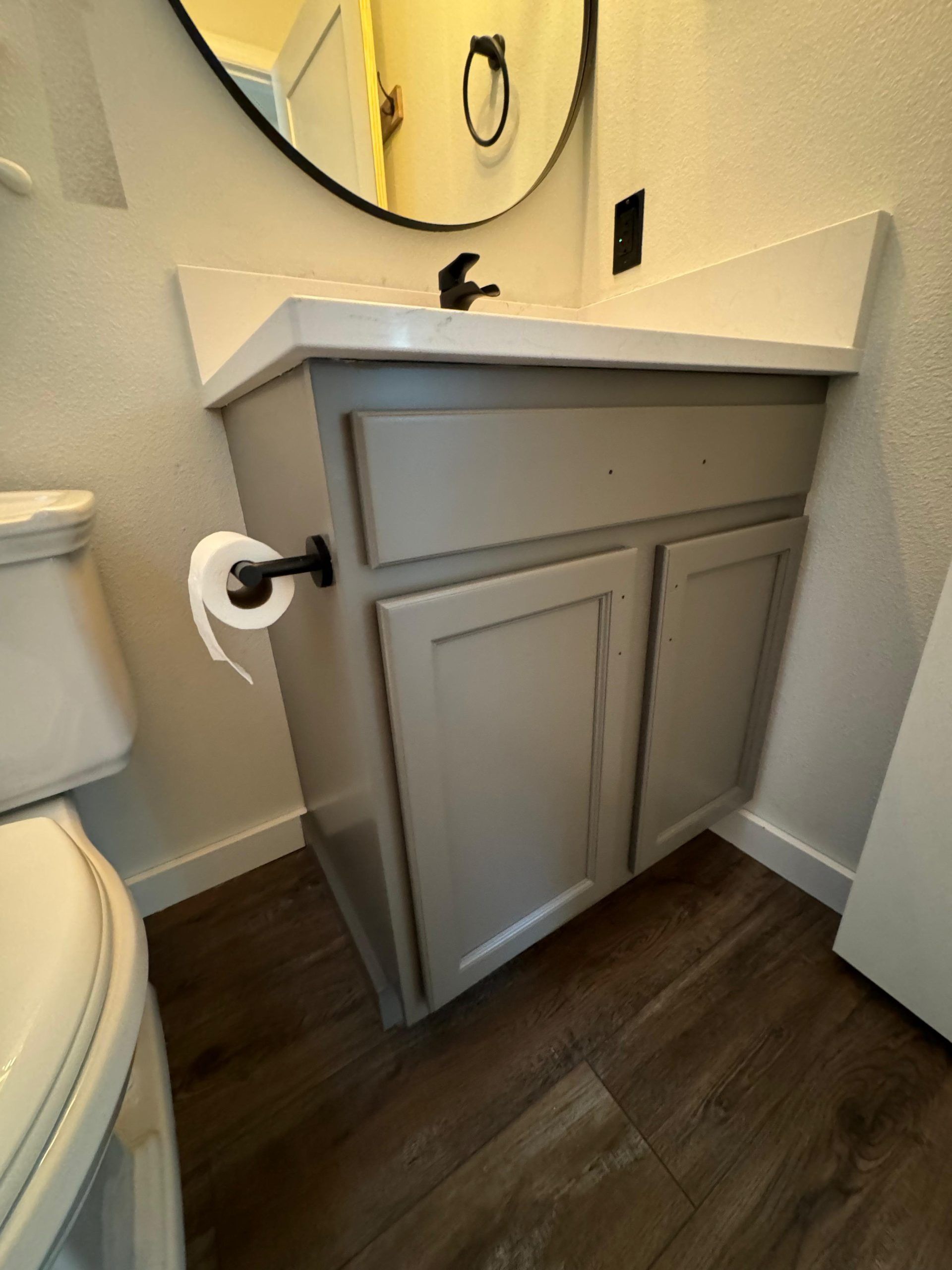 A gray bathroom vanity with a white countertop, circular mirror, and black hardware on a wood-look floor.