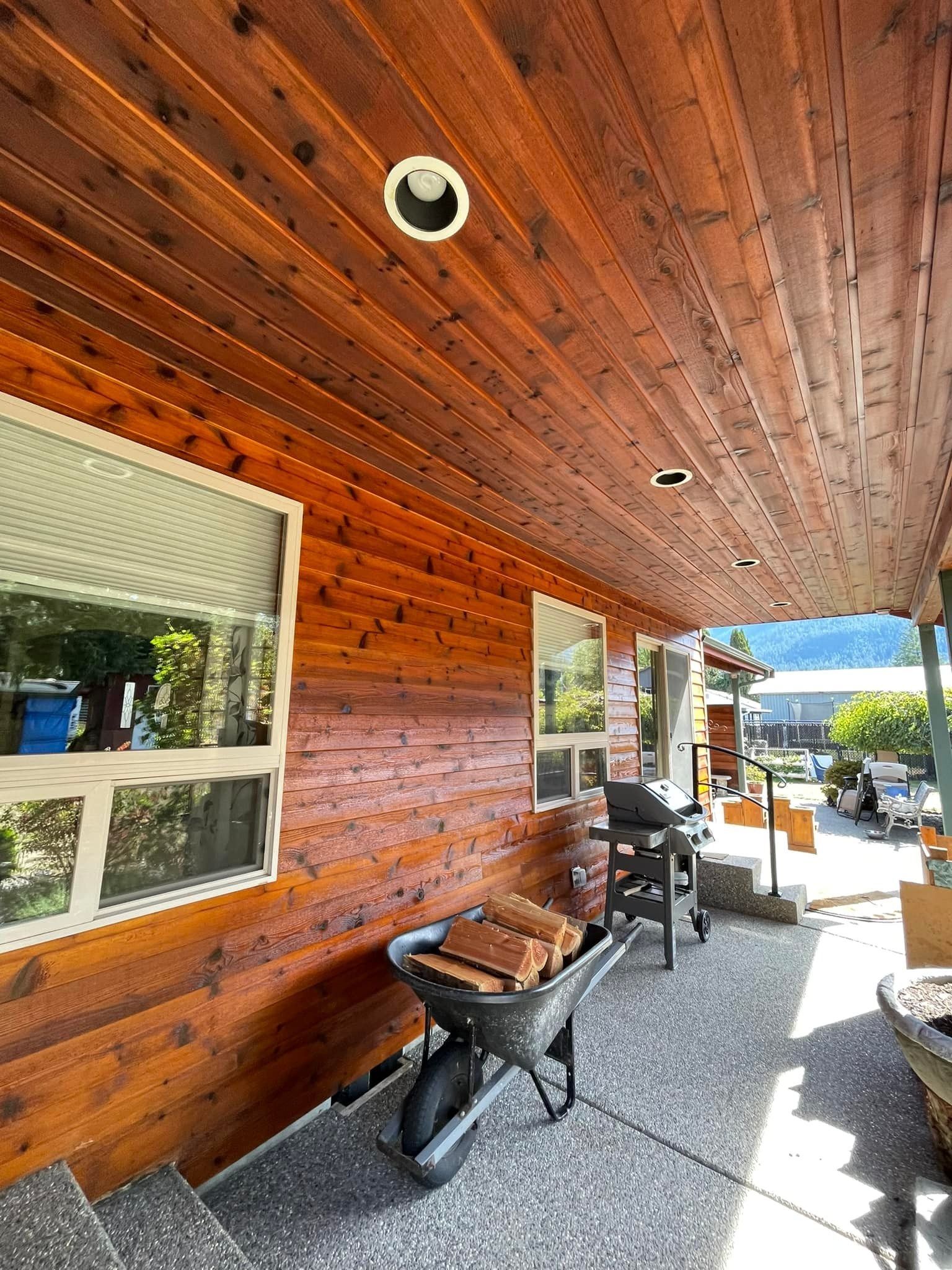 A covered patio with wood-paneled walls and ceilings, a wheelbarrow of firewood, and a grill on a pebble-textured floor.