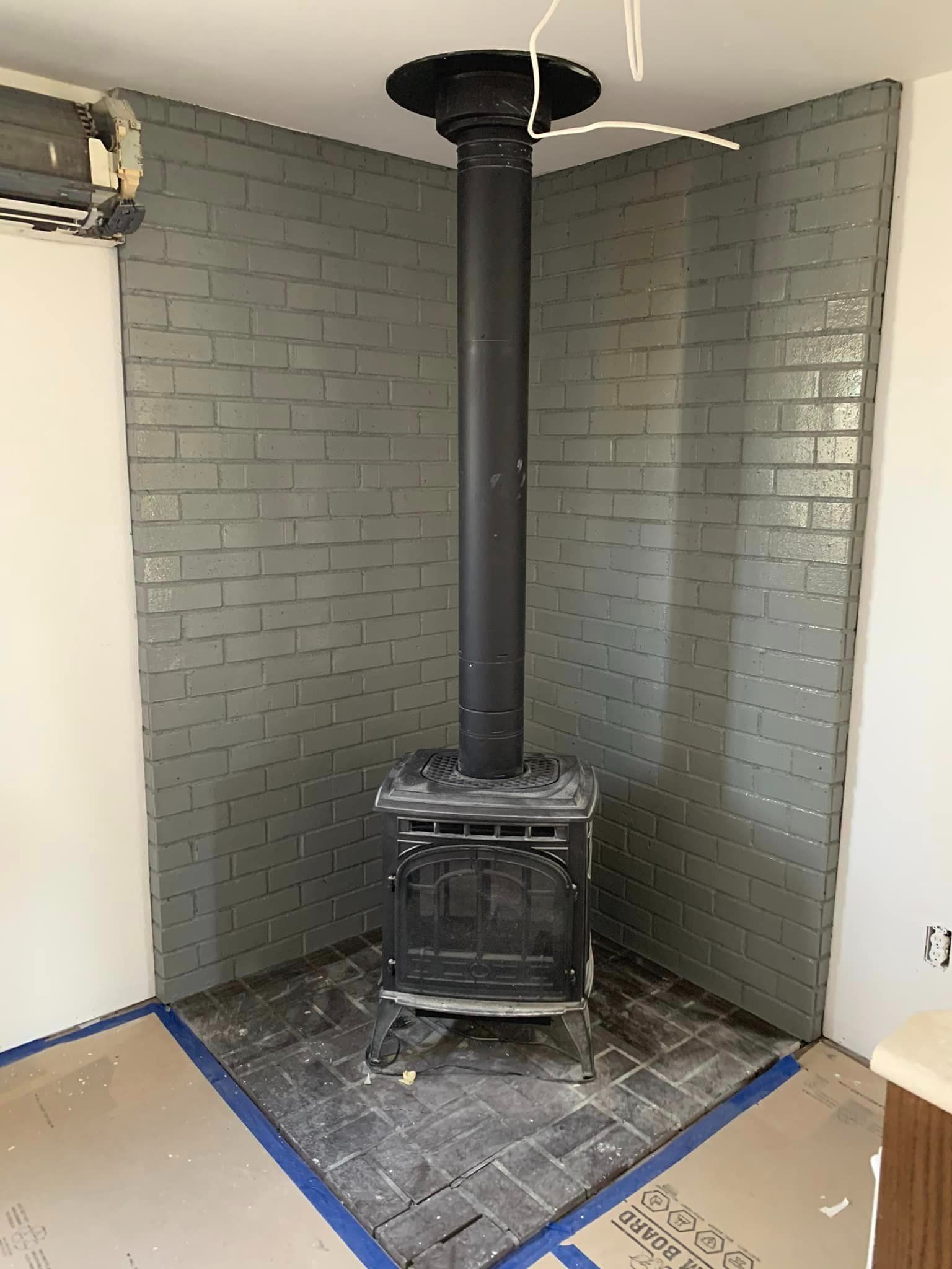 A black wood-burning stove stands on a grey tiled floor against a corner wall of grey-painted bricks, with a metal chimney.