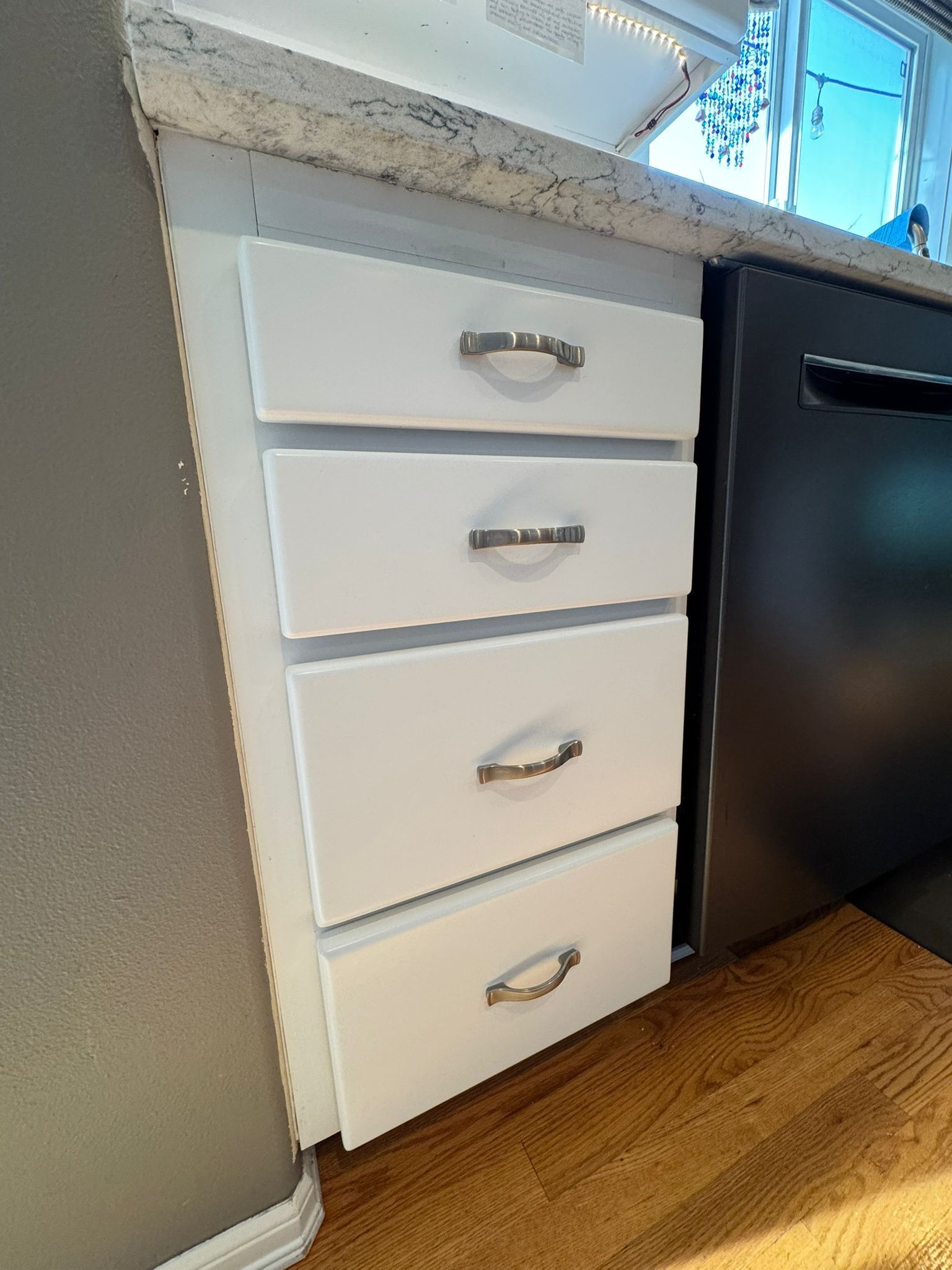 A stack of four white kitchen drawers with silver pulls, situated next to a black dishwasher under a granite countertop.