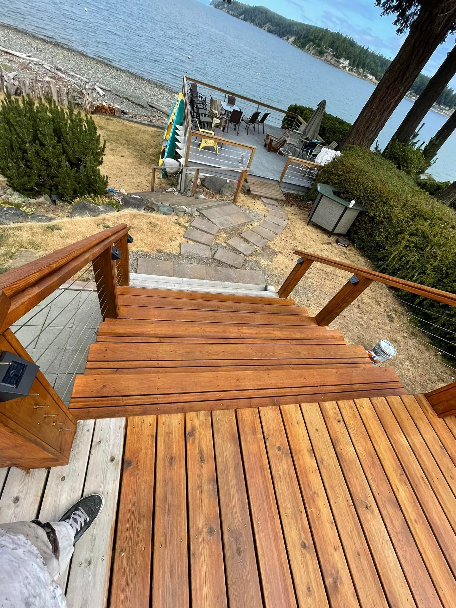 A high-angle view of wooden deck stairs leading down to a waterfront patio with a view of a lake and forest.
