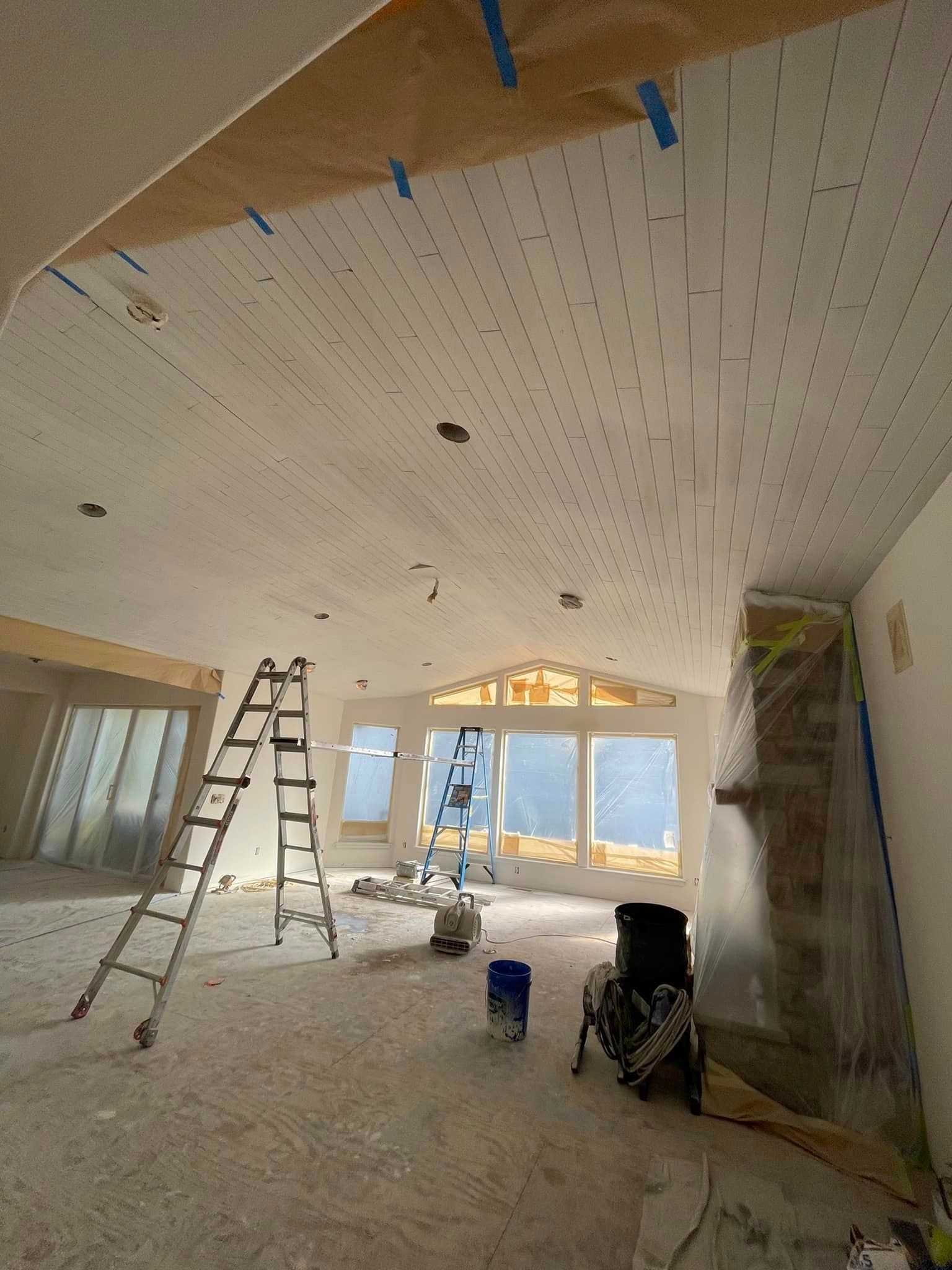 An interior room under construction features a vaulted ceiling with exposed screws, a tall ladder, and plastic-covered walls.