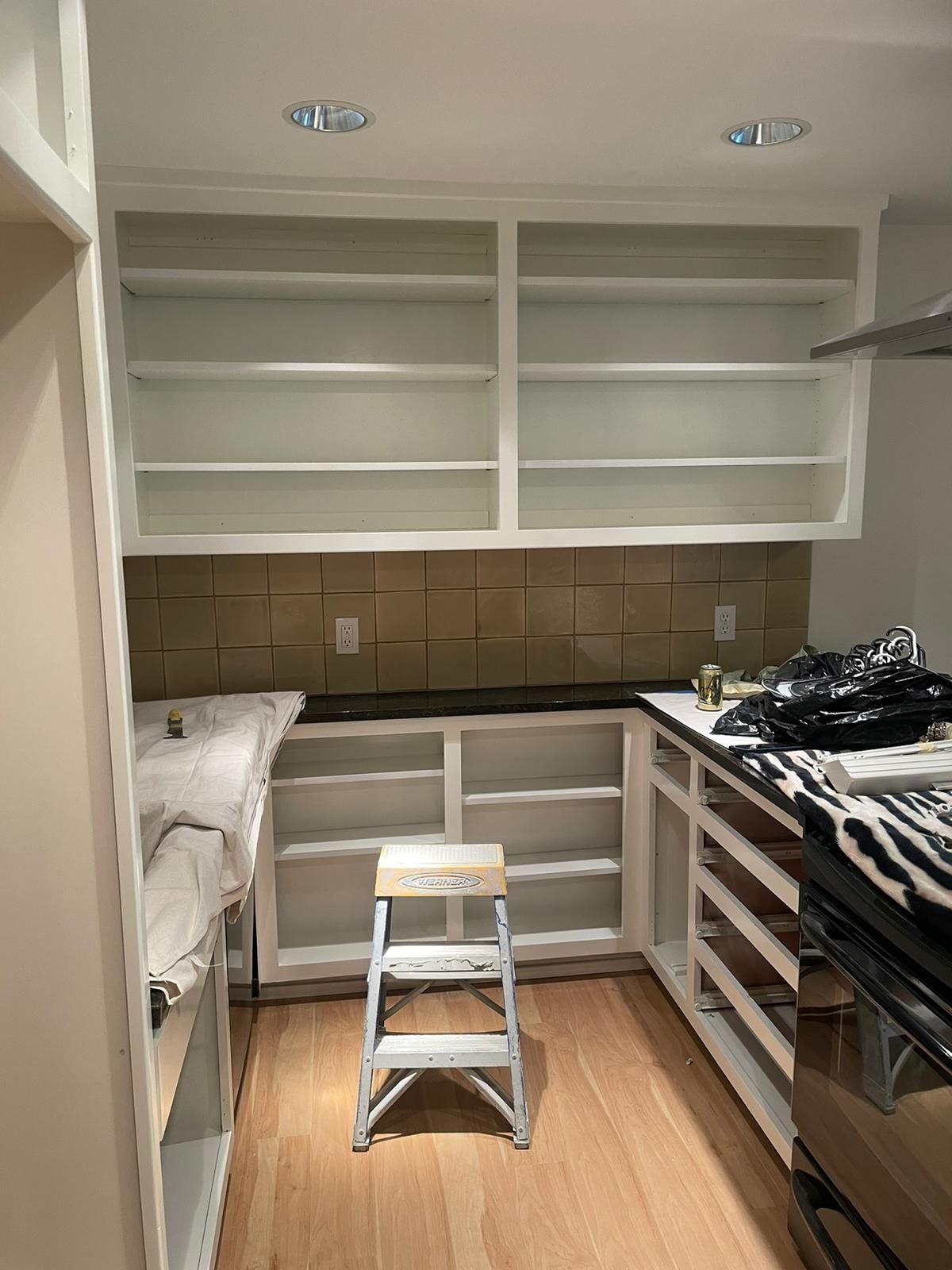 A room undergoing kitchen renovation featuring white cabinets, wood flooring, and a tan tile backsplash with a step stool.
