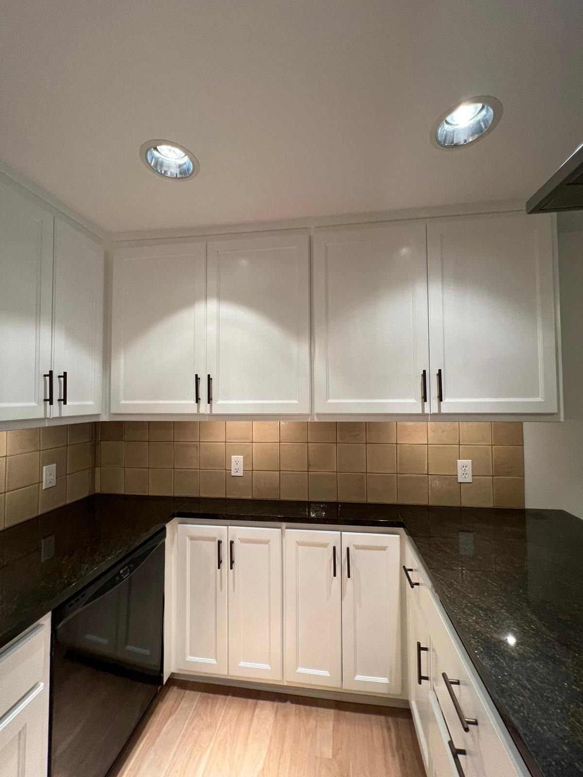 A kitchen with white cabinets, dark granite countertops, tan square tiled backsplash, and recessed ceiling lights.