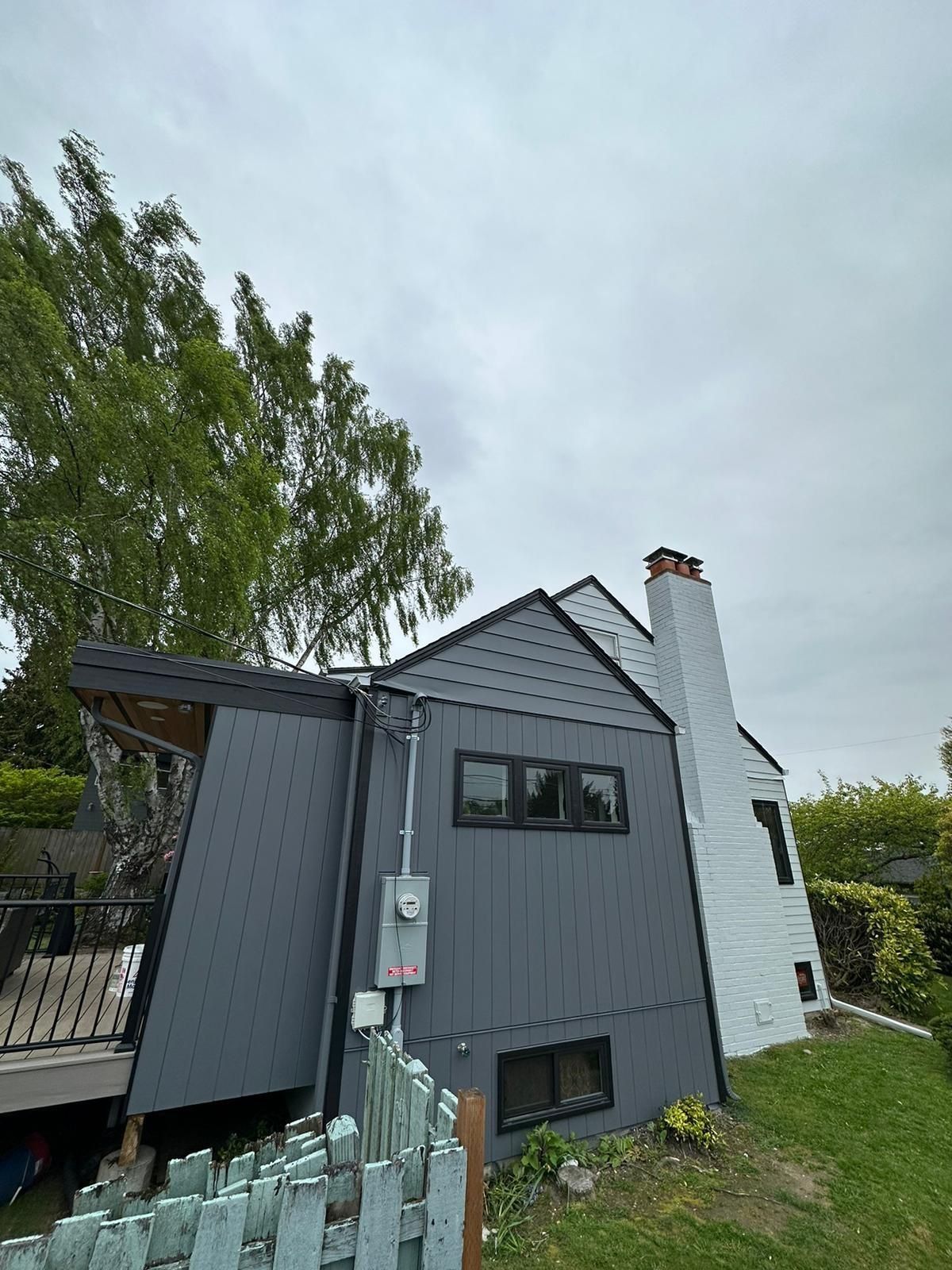 A gray, modern-sided home addition attaches to a white brick house with a chimney under a cloudy sky.