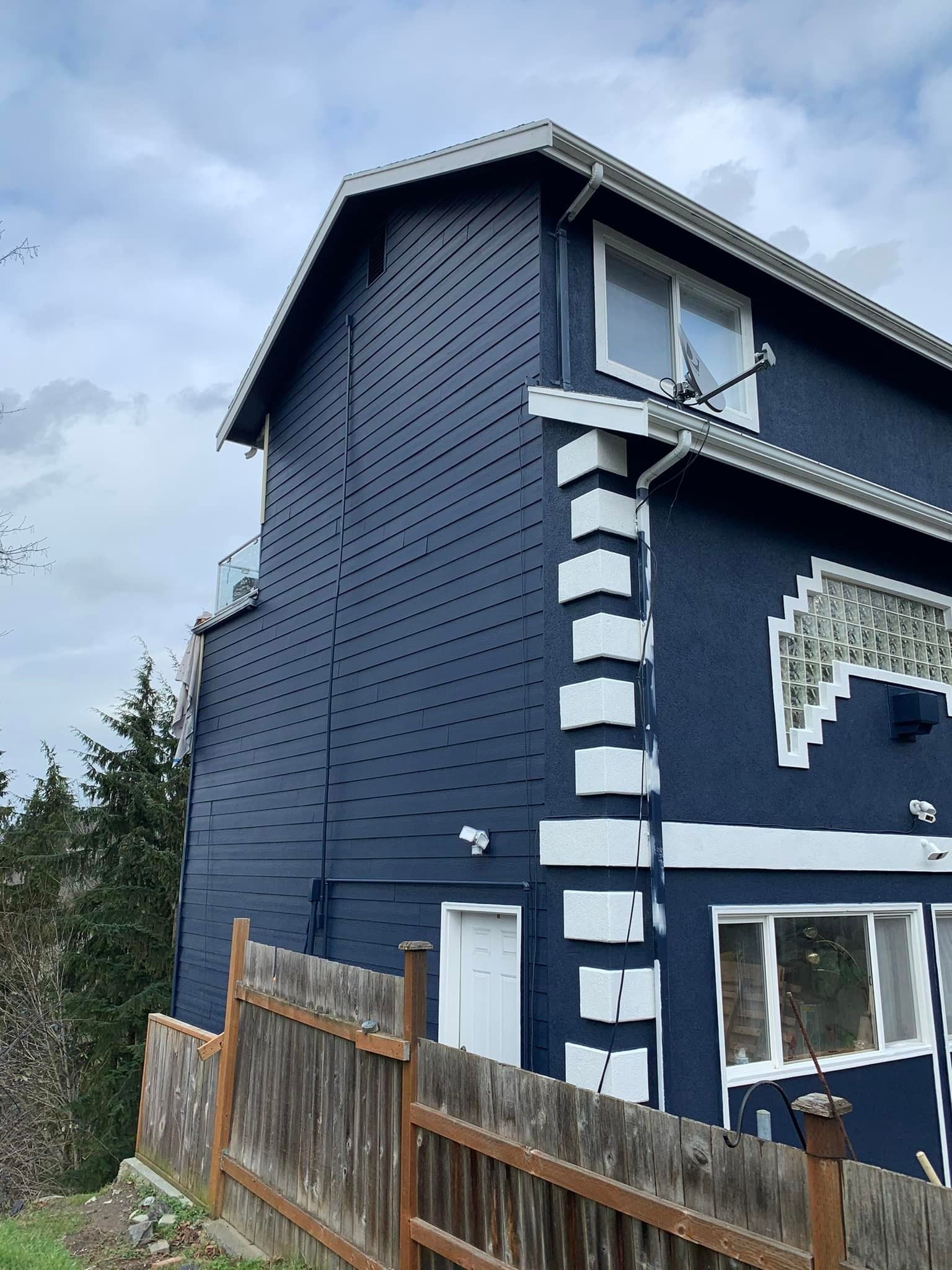 A two-story blue house with white brick-patterned corner accents, viewed from the side behind a wooden fence.