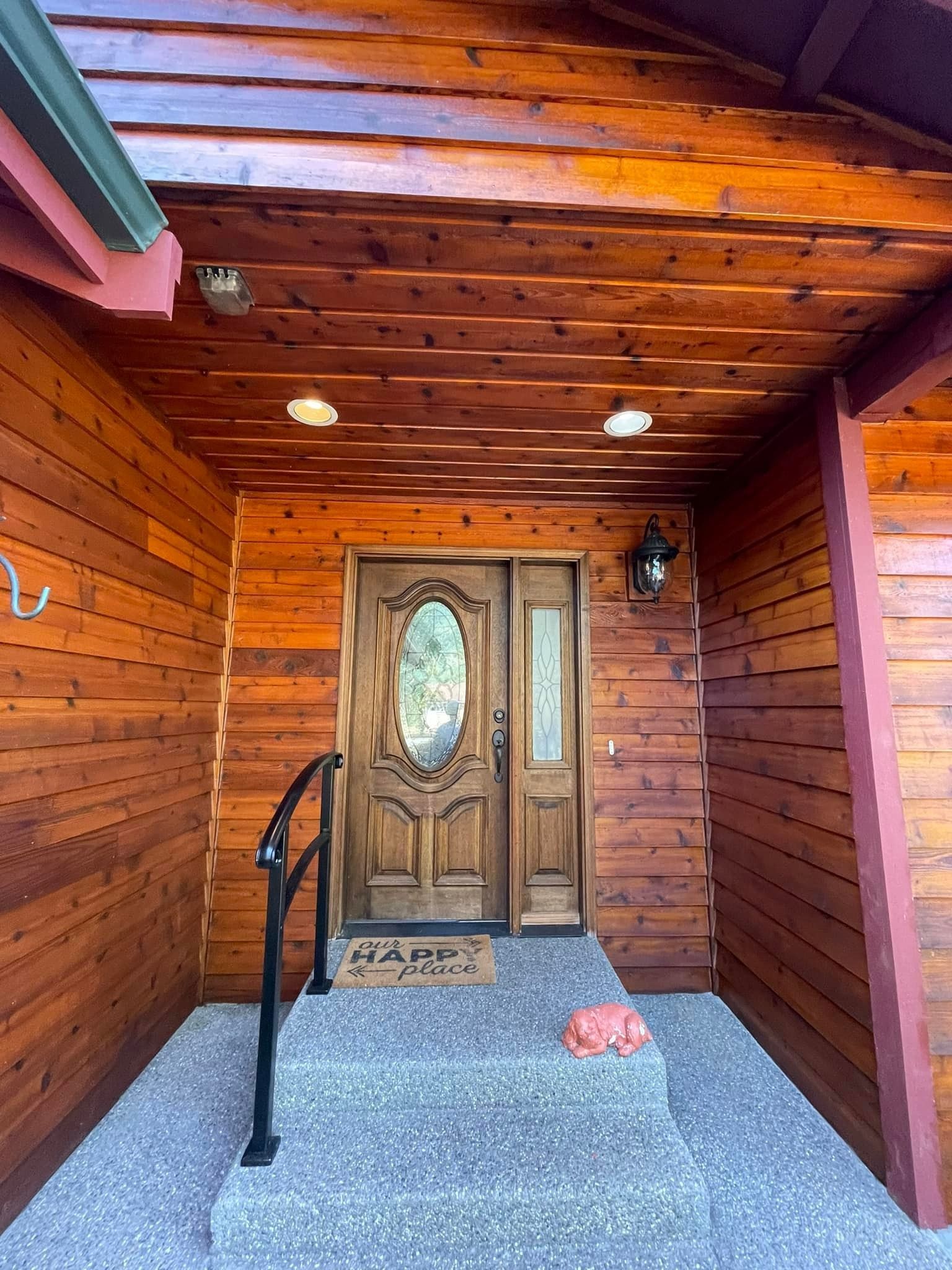 A wooden porch with a gravel landing, a black metal handrail, and a decorative wooden front door under a roof overhang.