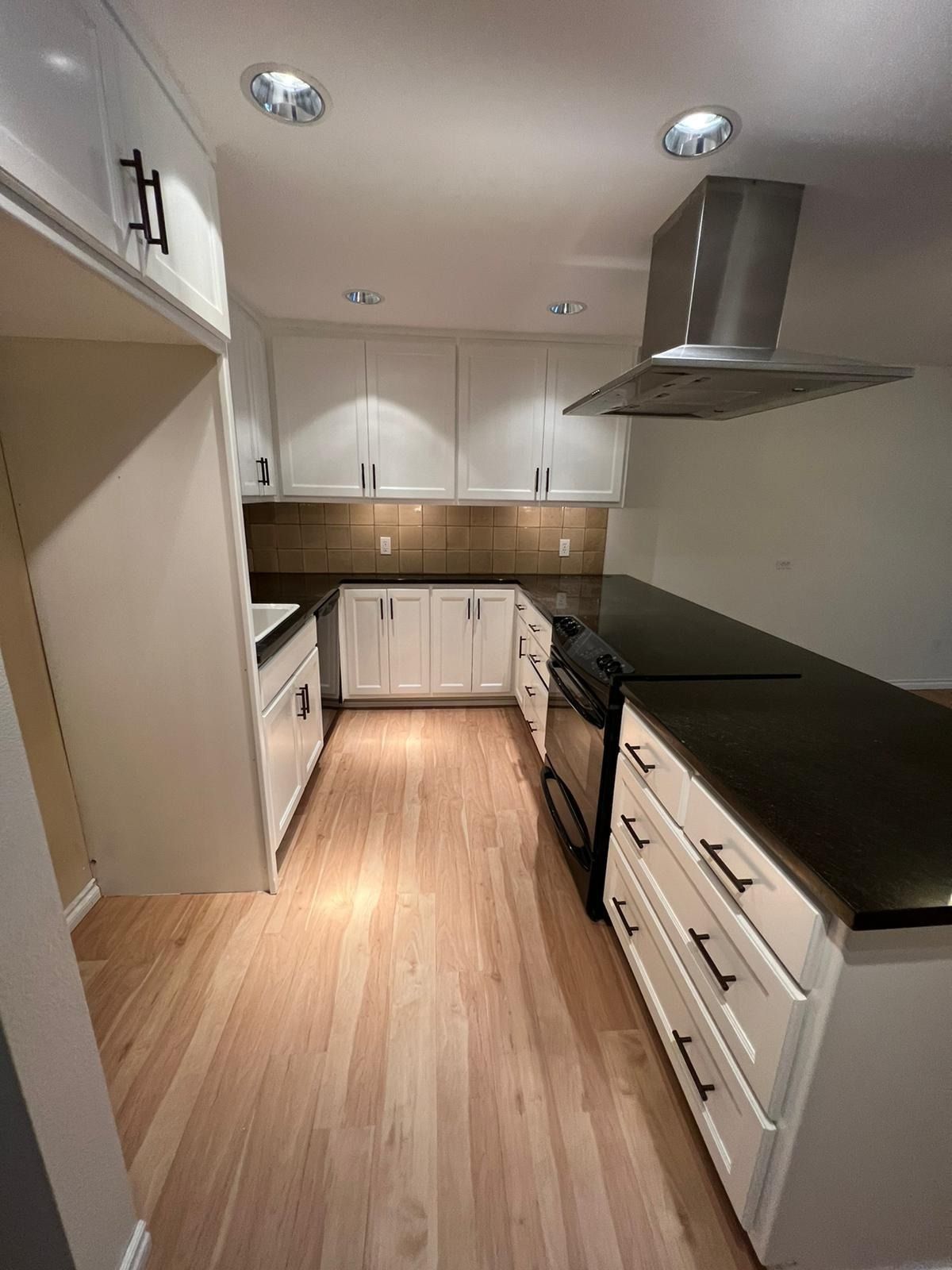 A modern kitchen with white cabinets, dark countertops, a stainless steel range hood, and light wood flooring.