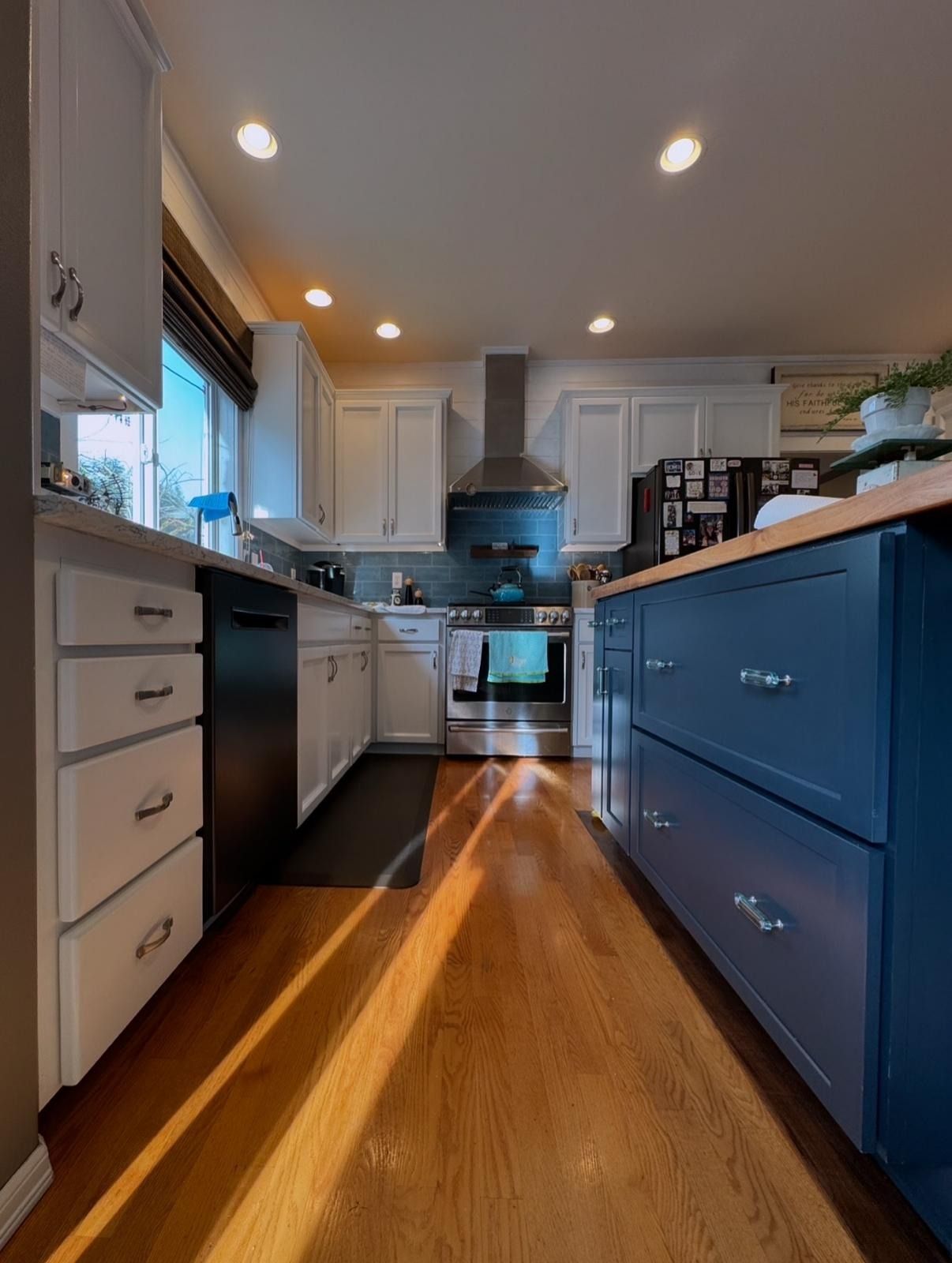 A modern kitchen with white upper cabinets, a dark blue island, stainless steel appliances, and wood floors with sunlight.
