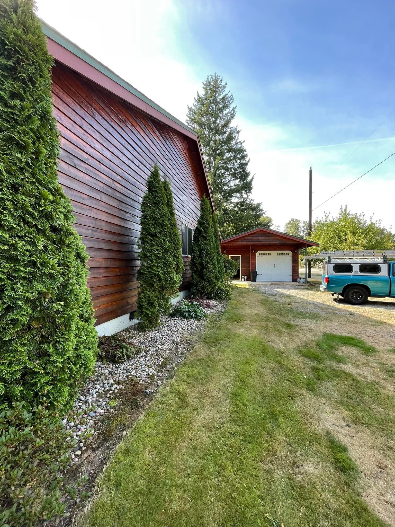 A view of a reddish-brown wooden building with evergreen bushes, leading to a detached garage and a blue pickup truck.