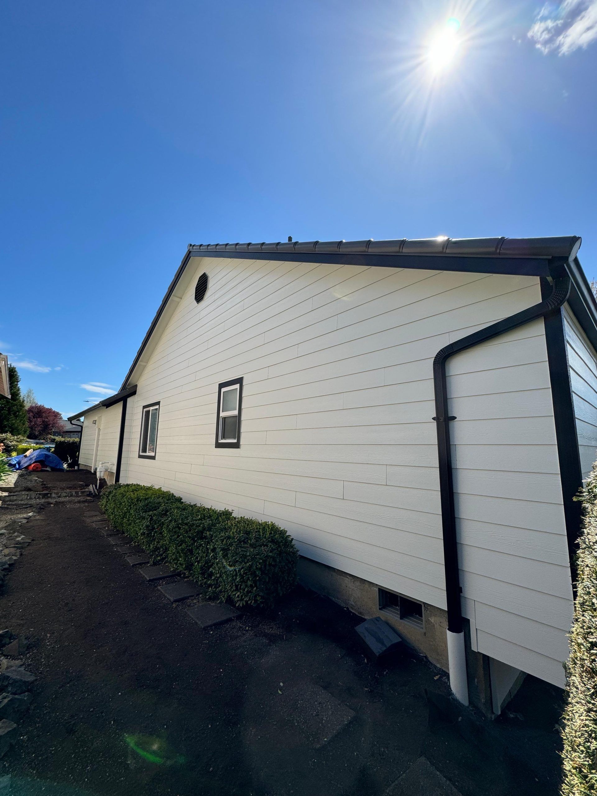A side view of a white, single-story house with dark trim, featuring two small windows and a hedge along the foundation.