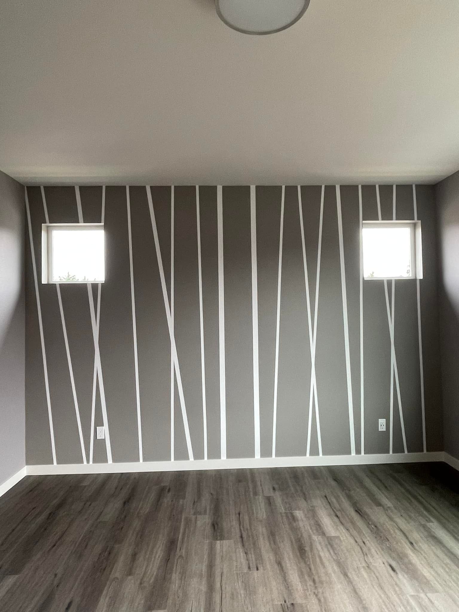 A room wall painted gray with vertical white stripes of varying widths and angles, flanked by two square windows.