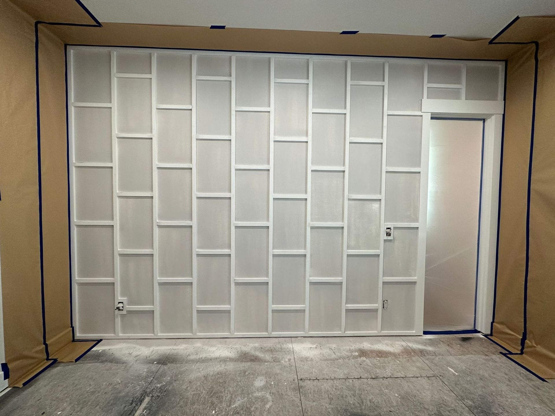 A white wall features a modern, vertical grid pattern of trim molding, with masking paper surrounding the work area.