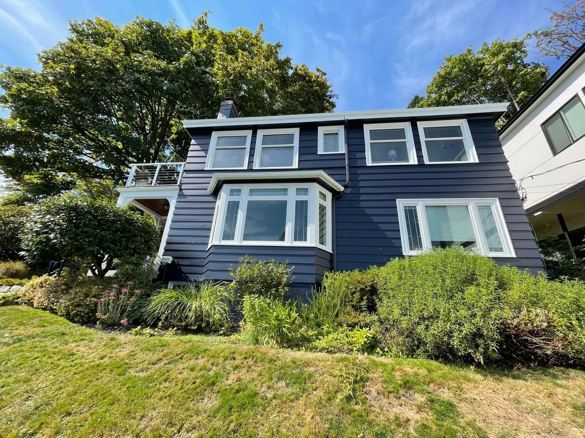 A two-story, dark blue house with white trim stands on a grassy hill under a bright blue sky.