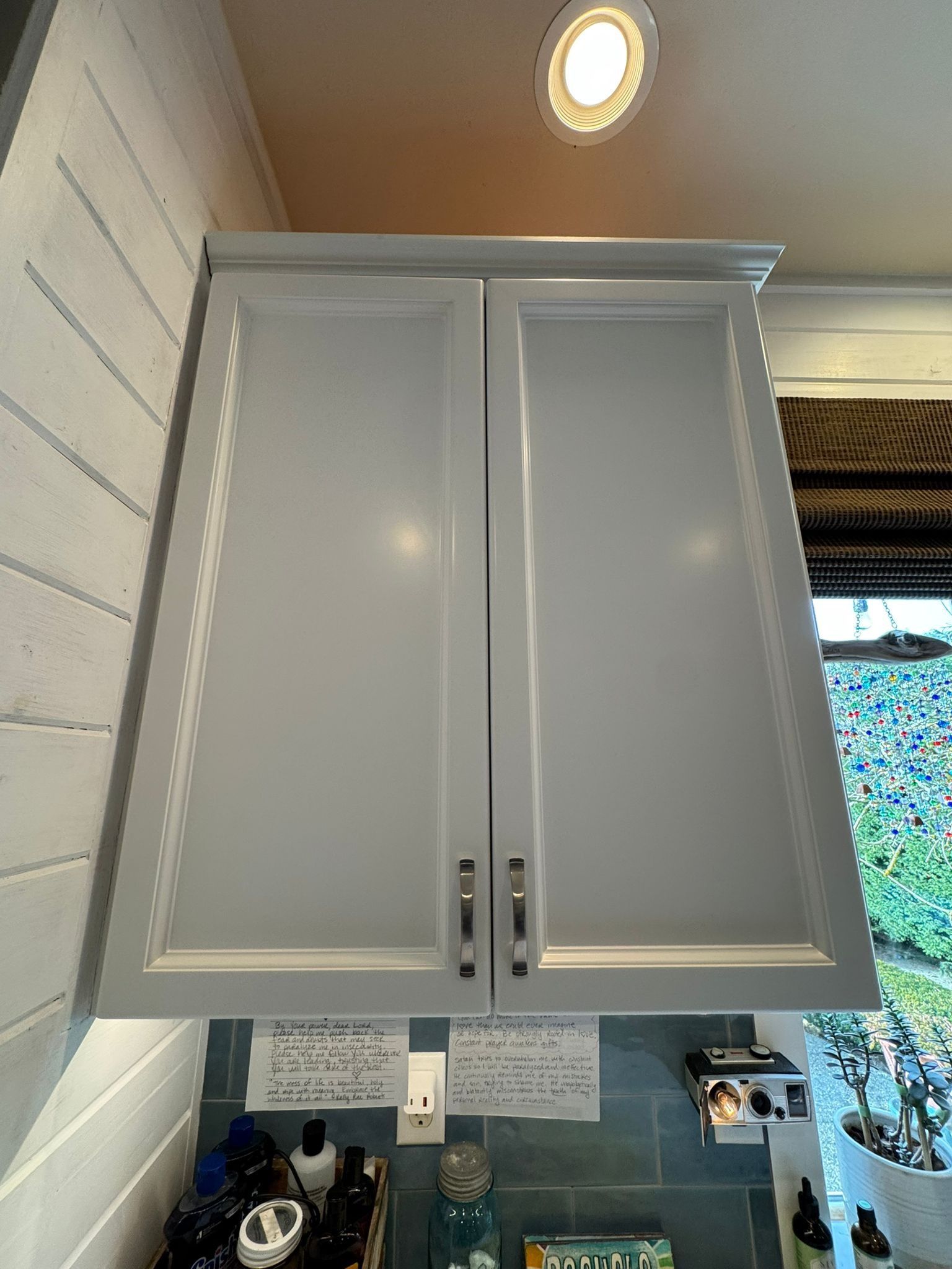 Two white Shaker-style kitchen cabinets mounted on a wall above a counter with various items and a window.