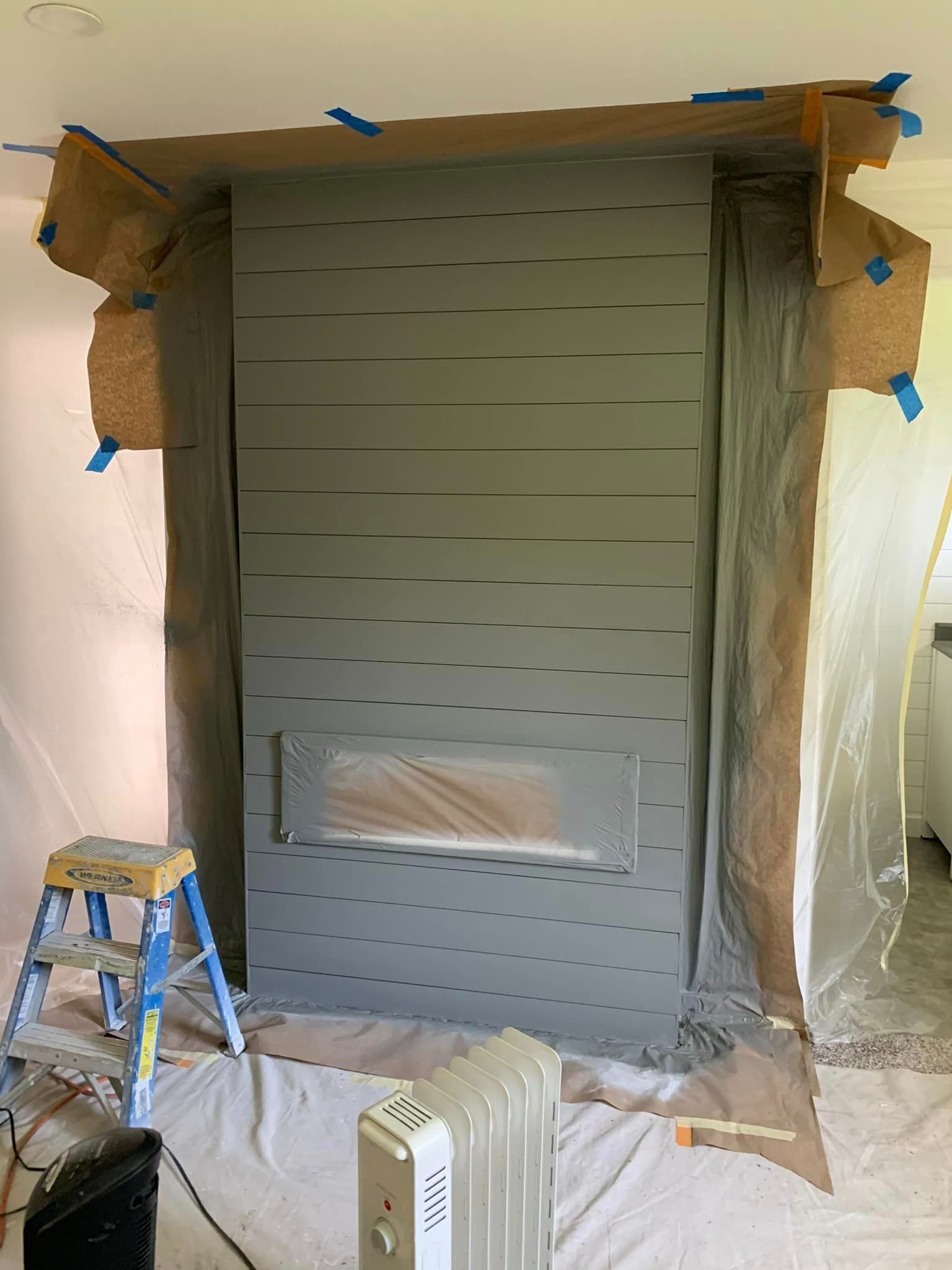 A fireplace wall being painted grey, with surrounding areas masked off with paper and plastic, and a ladder nearby.