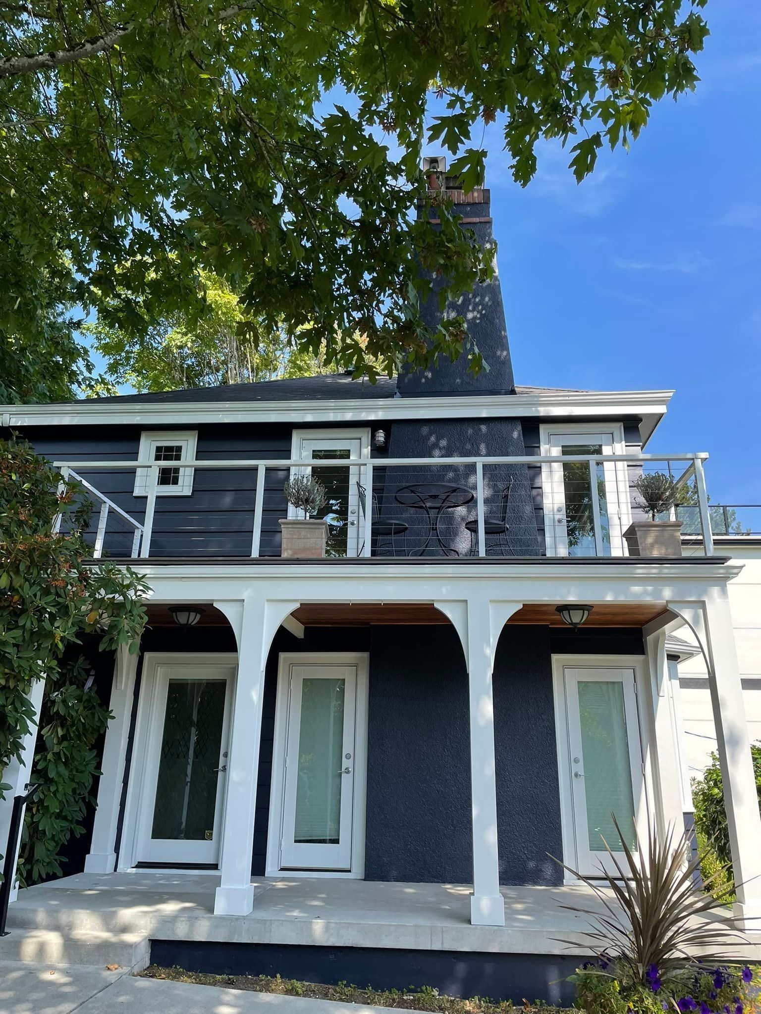 A two-story dark-painted house with a white wraparound porch, a second-floor balcony, and a prominent brick chimney.