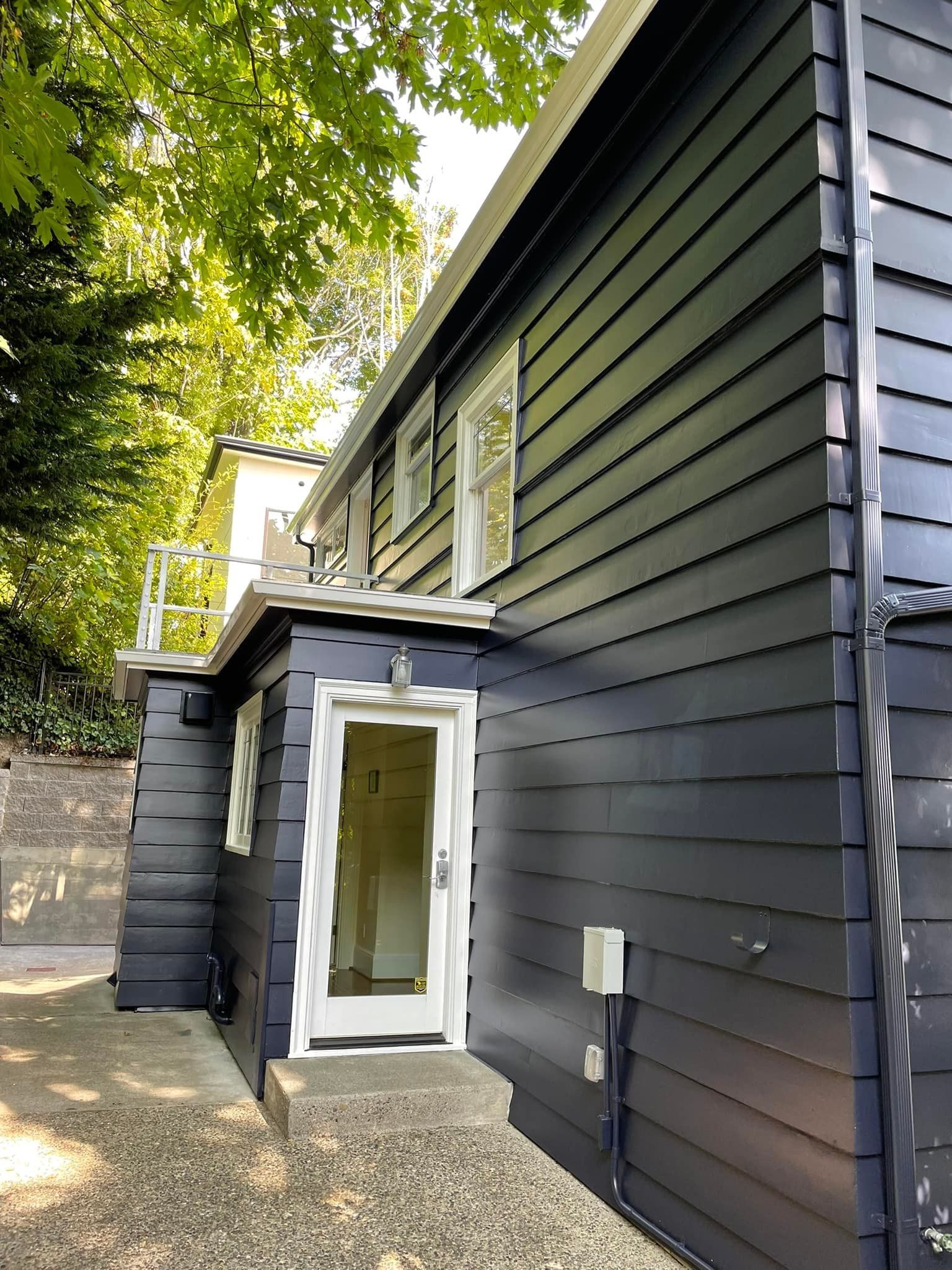 A dark-painted wooden house exterior featuring a side entrance door with white trim and stone gravel ground cover.