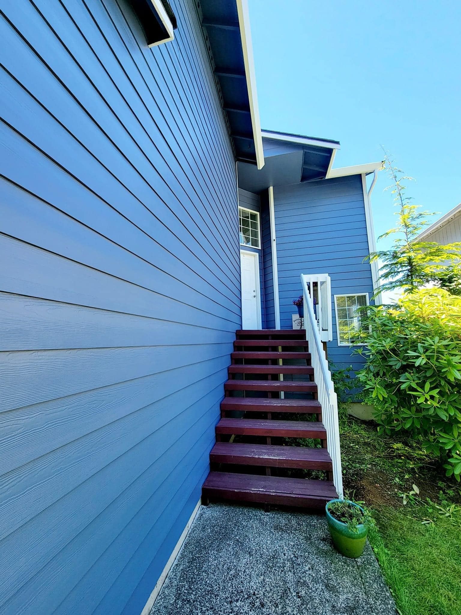 A wooden staircase with dark brown steps leads up to a side door of a blue-sided house with a clear blue sky above.