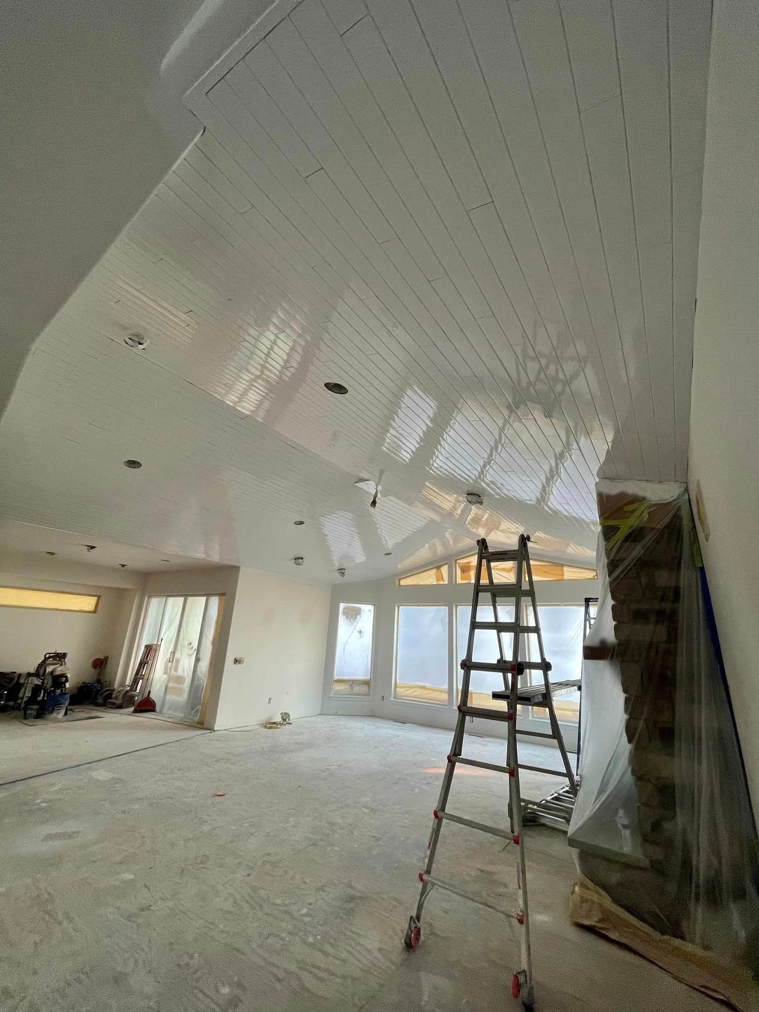 An unfinished room under construction featuring a vaulted white paneled ceiling, a metal ladder, and large windows.
