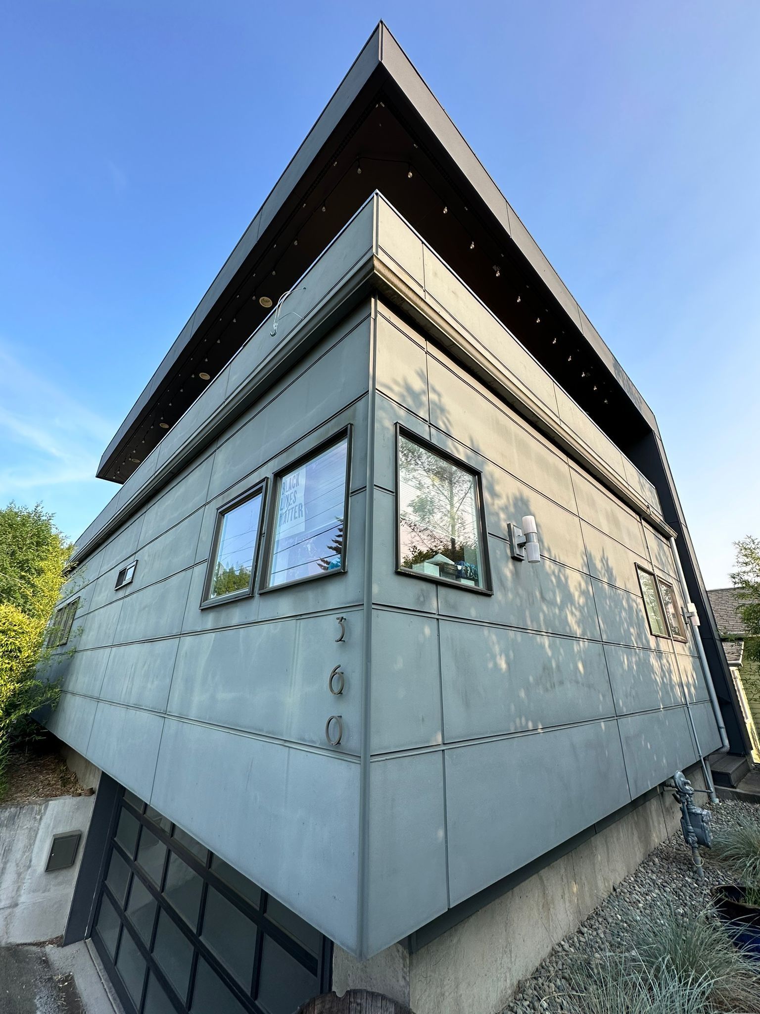 Low-angle view of a modern house with gray paneled siding, a dark roof overhang, and a garage, set against a blue sky.