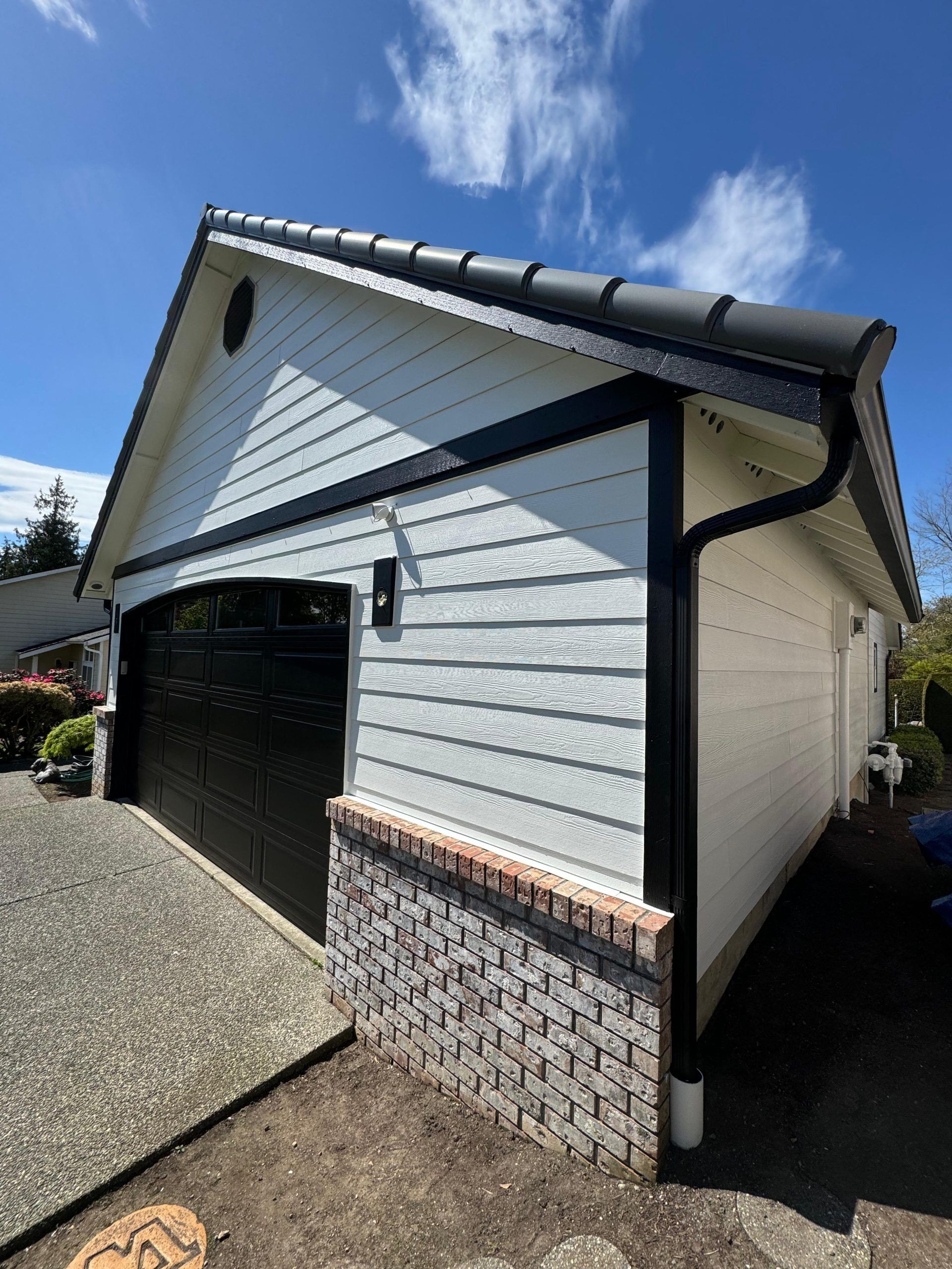 White detached garage with black trim, a black garage door, and a brick base under a bright blue sky.