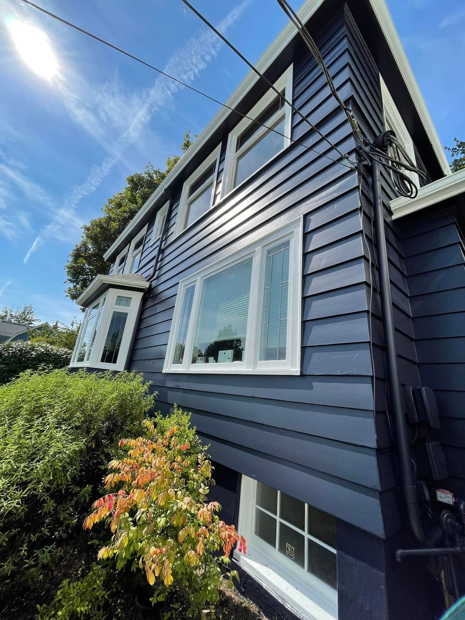 A low-angle view of a dark blue, wood-sided house with white window frames under a sunny blue sky.