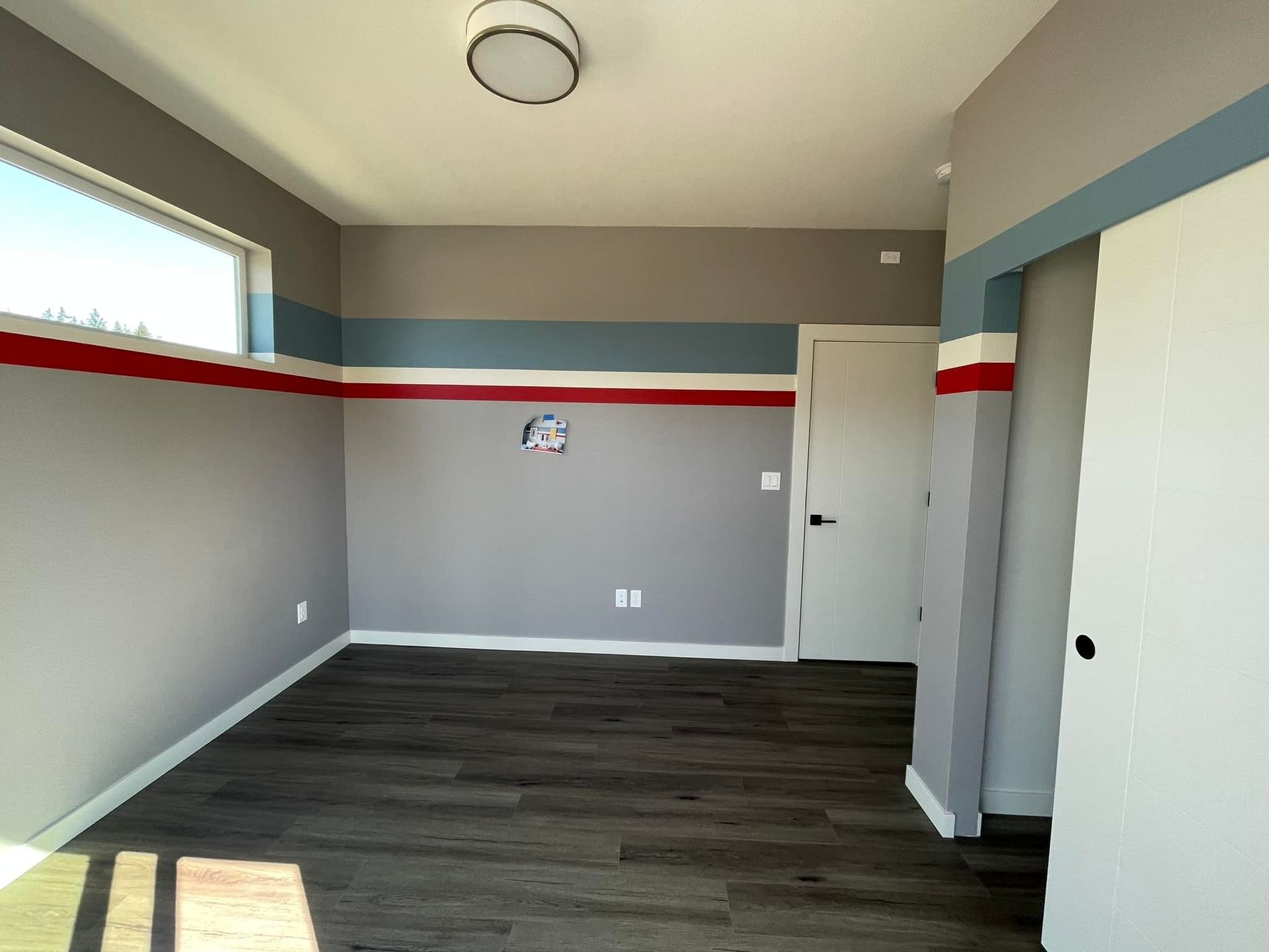 An empty room with gray walls featuring a horizontal blue and red striped accent, white trim, and dark wood flooring.