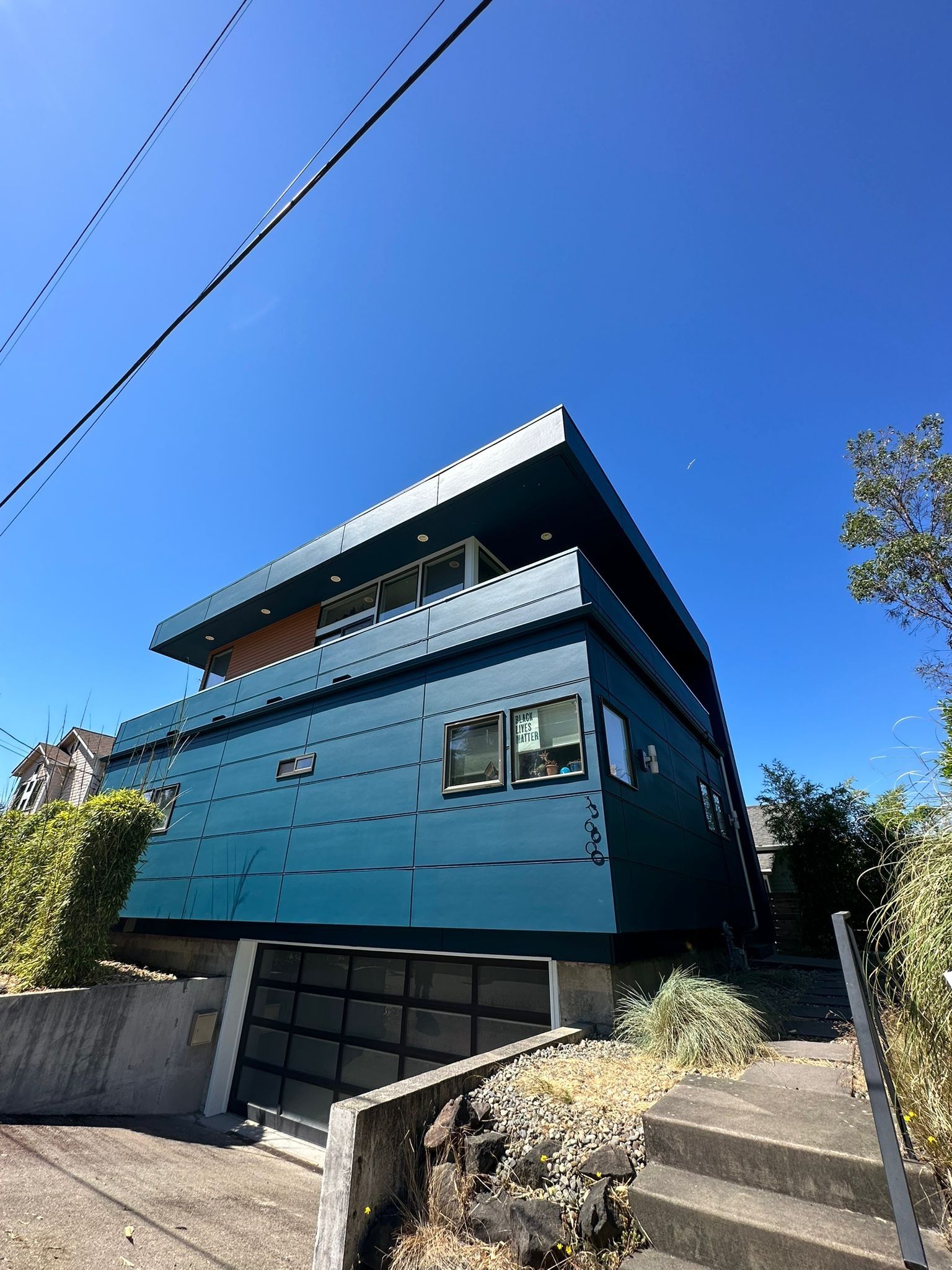 A modern, blue two-story house with horizontal paneling and a dark garage door, set against a bright, clear blue sky.