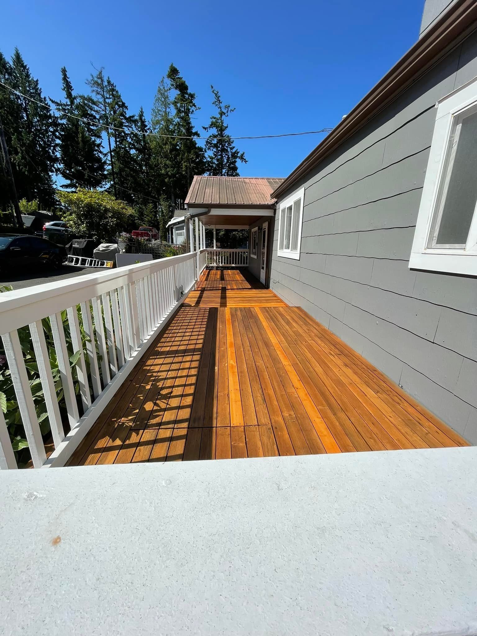 A sunlit wooden deck runs alongside a grey house with a white railing and a covered porch under a clear blue sky.