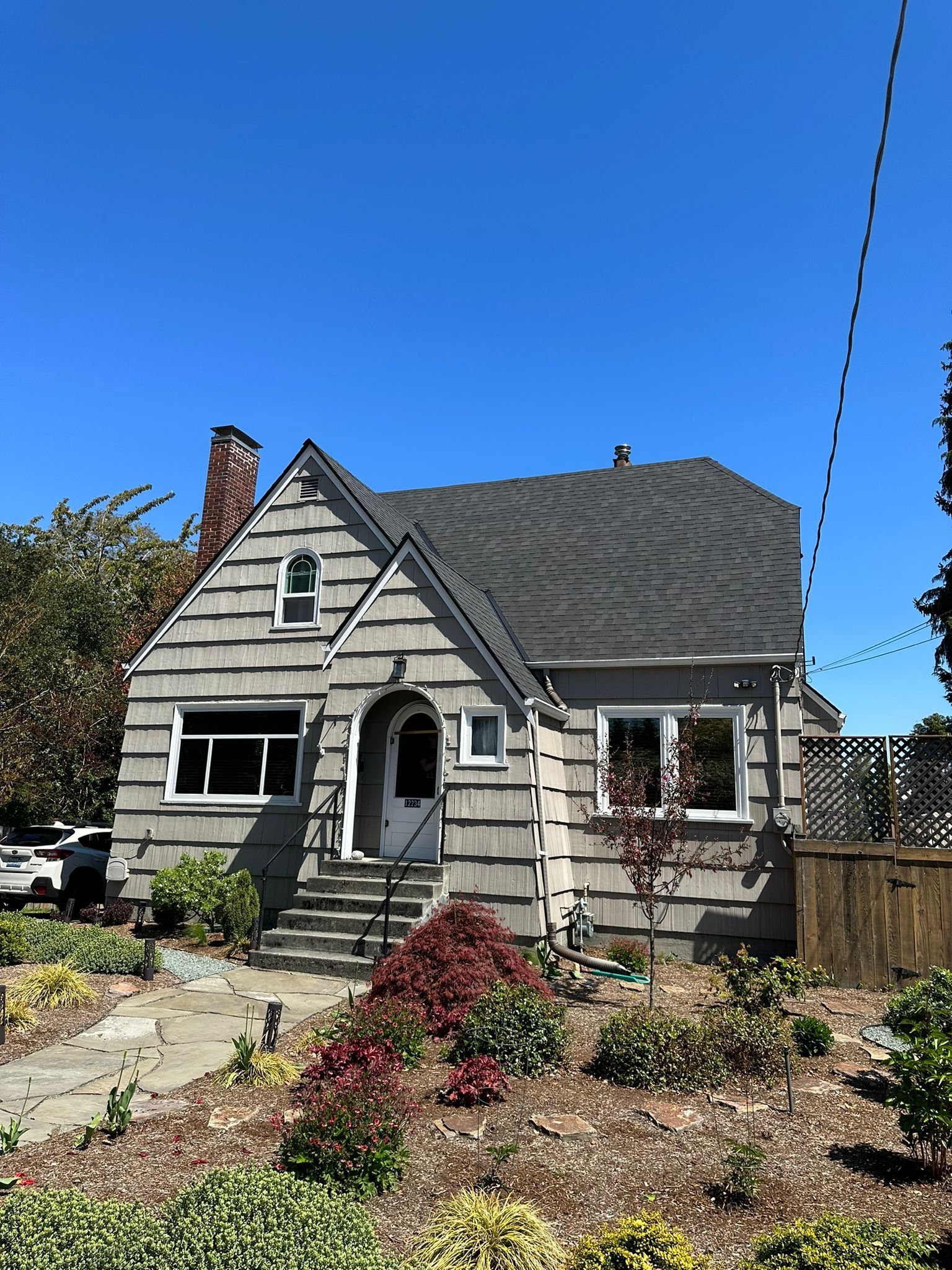 A one-story grey house with a steeply pitched roof, a red brick chimney, and a small front yard under a clear blue sky.