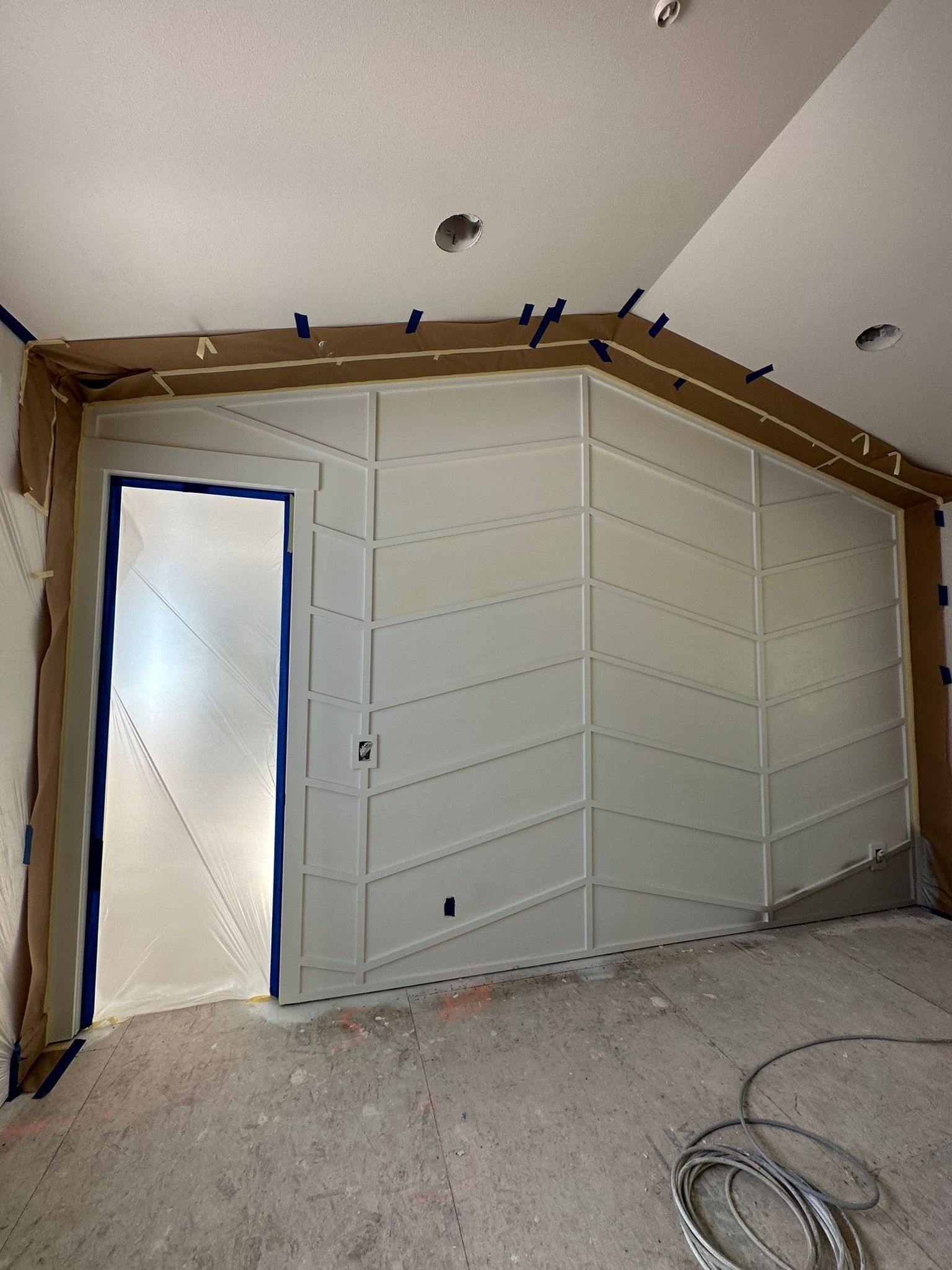 A wall in a room under construction features a chevron-patterned wood trim design and a door covered in plastic sheeting.