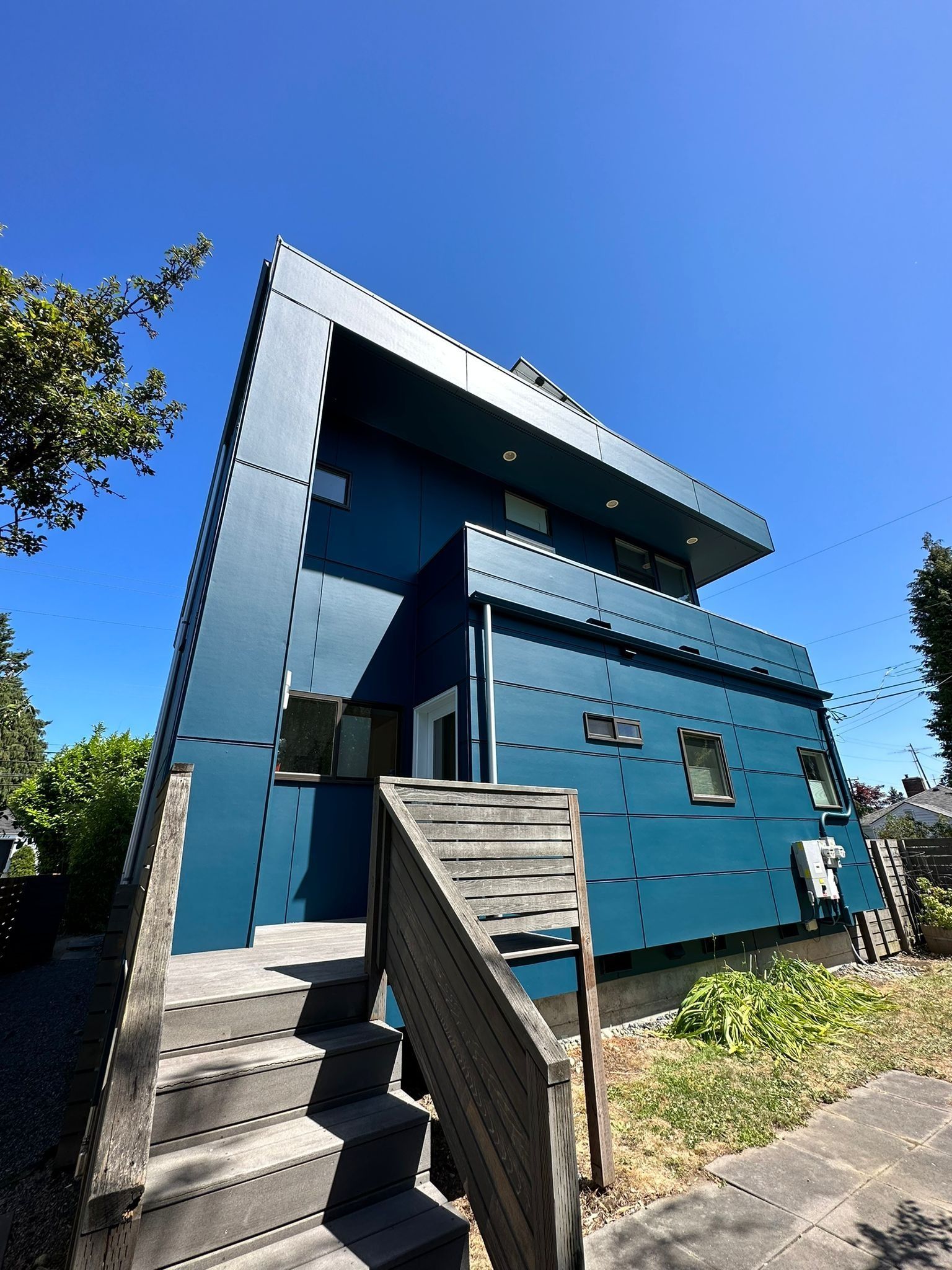 A low-angle view of a modern two-story house with dark blue exterior panels, a concrete staircase, and a clear blue sky.