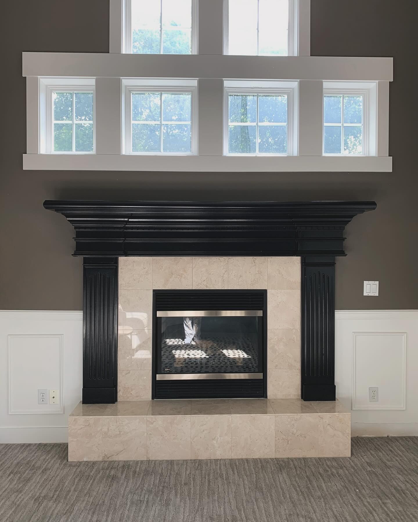 A black mantel fireplace with beige tile surround, set against a brown wall with white wainscoting and upper windows.