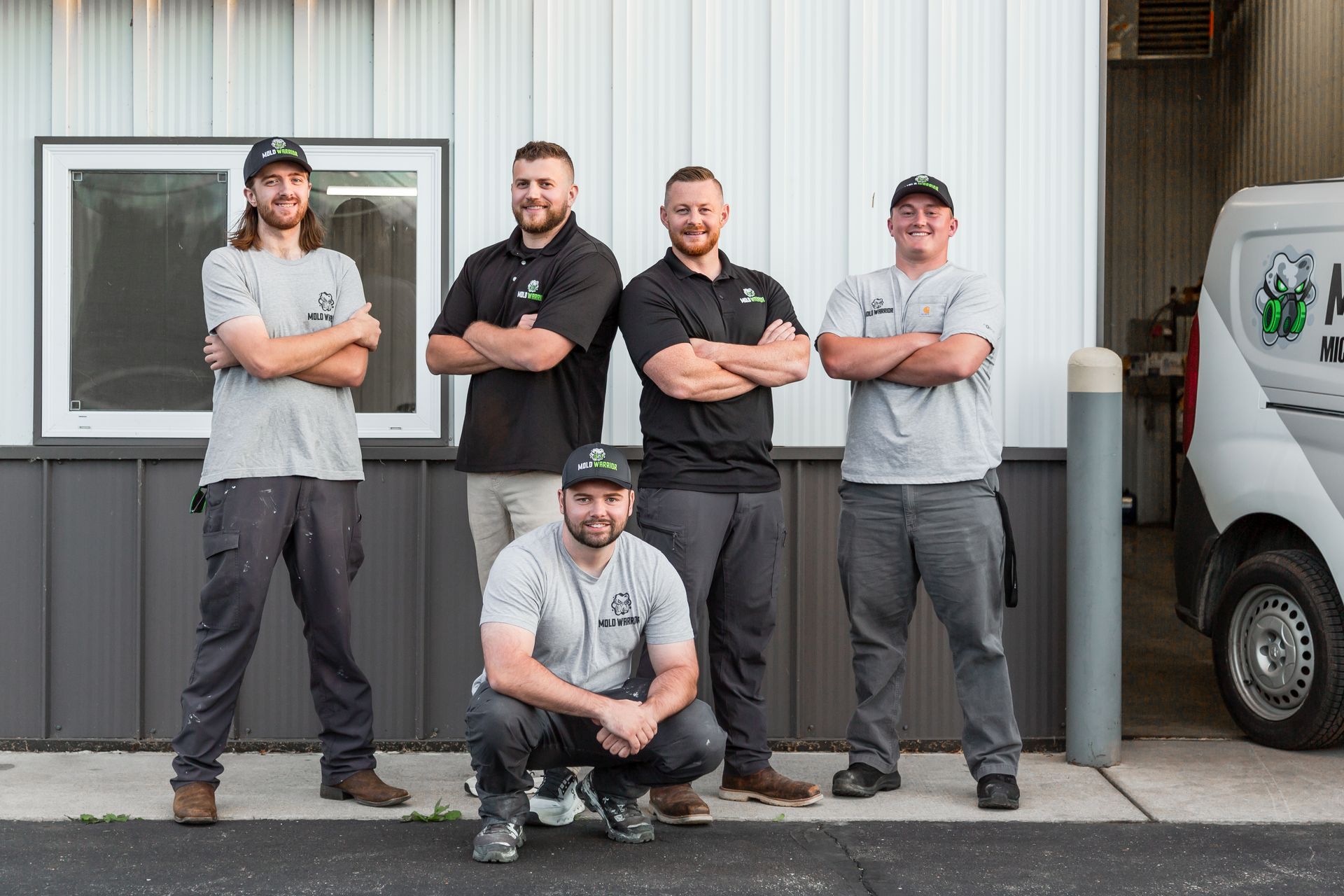 Five men, arms crossed, pose in front of a building. One crouches. Building has a window and a van.