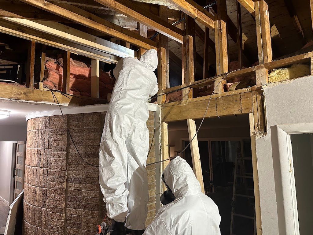 Two workers in protective suits removing insulation from a wooden wall.