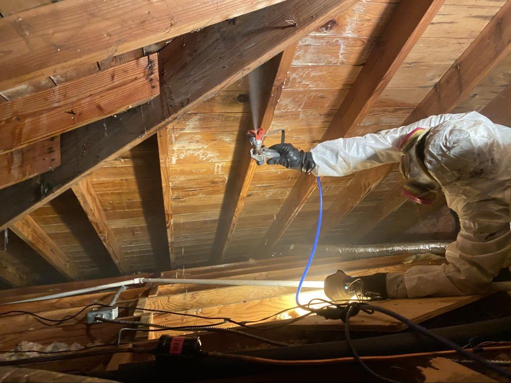 Person in protective suit sprays insulation in an attic, brown wooden beams overhead.