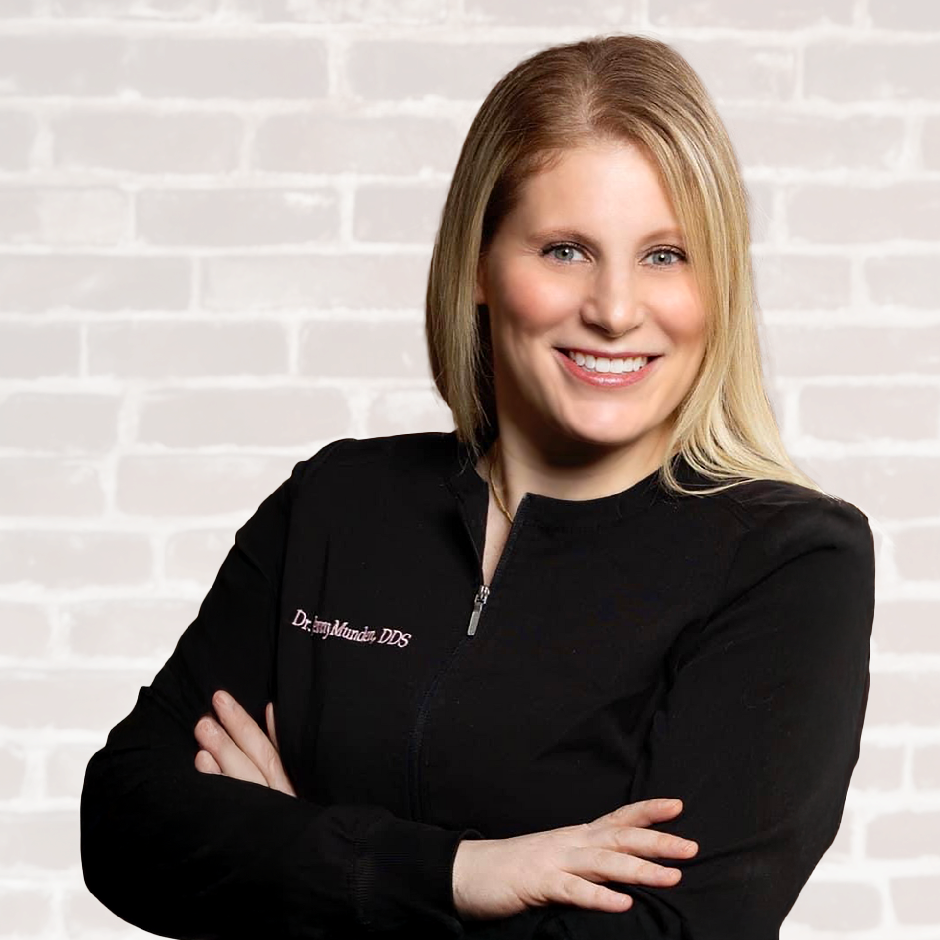 Blonde woman with arms crossed, smiling, wearing a black jacket against a white brick wall.