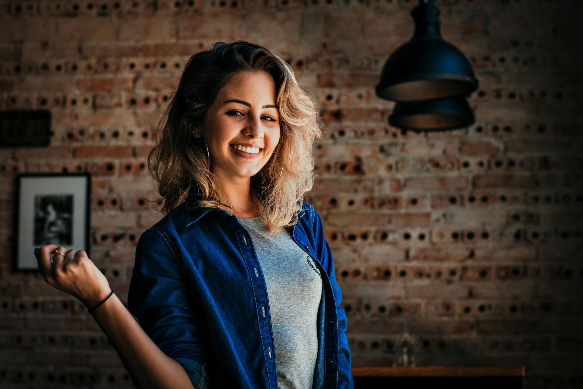 Smiling woman in a blue denim shirt and gray top, in front of a brick wall and hanging lamp.