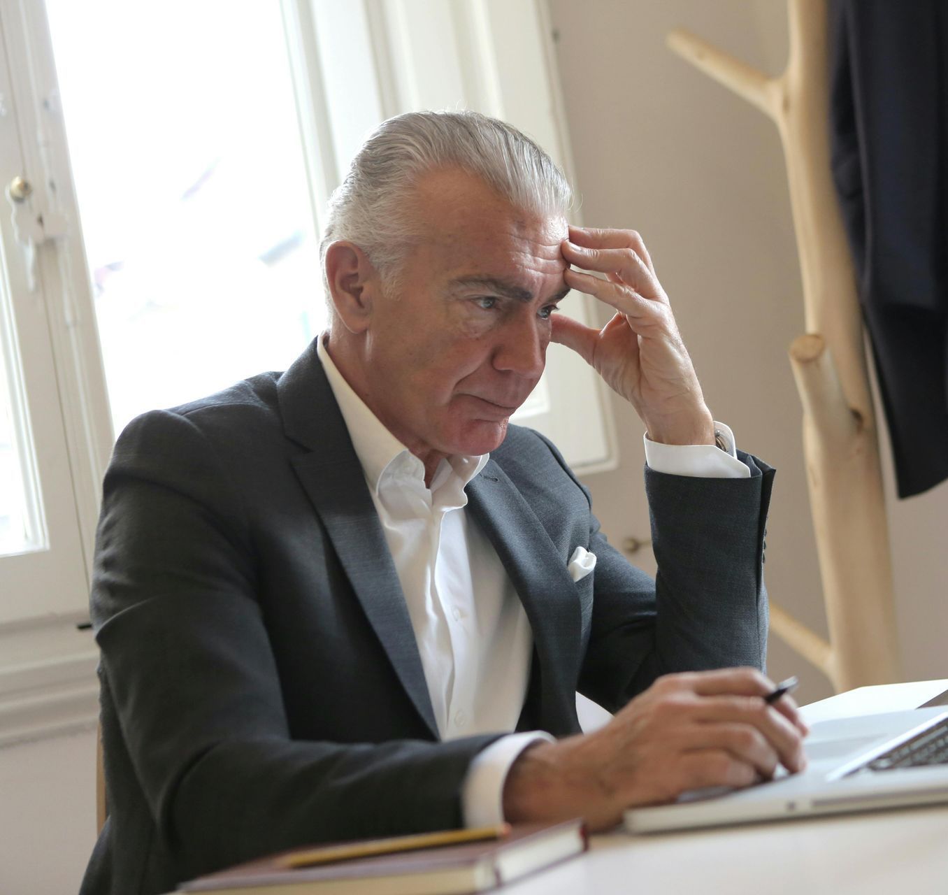 Man in a suit looking stressed, holding his head, and working on a laptop at a desk near a window.