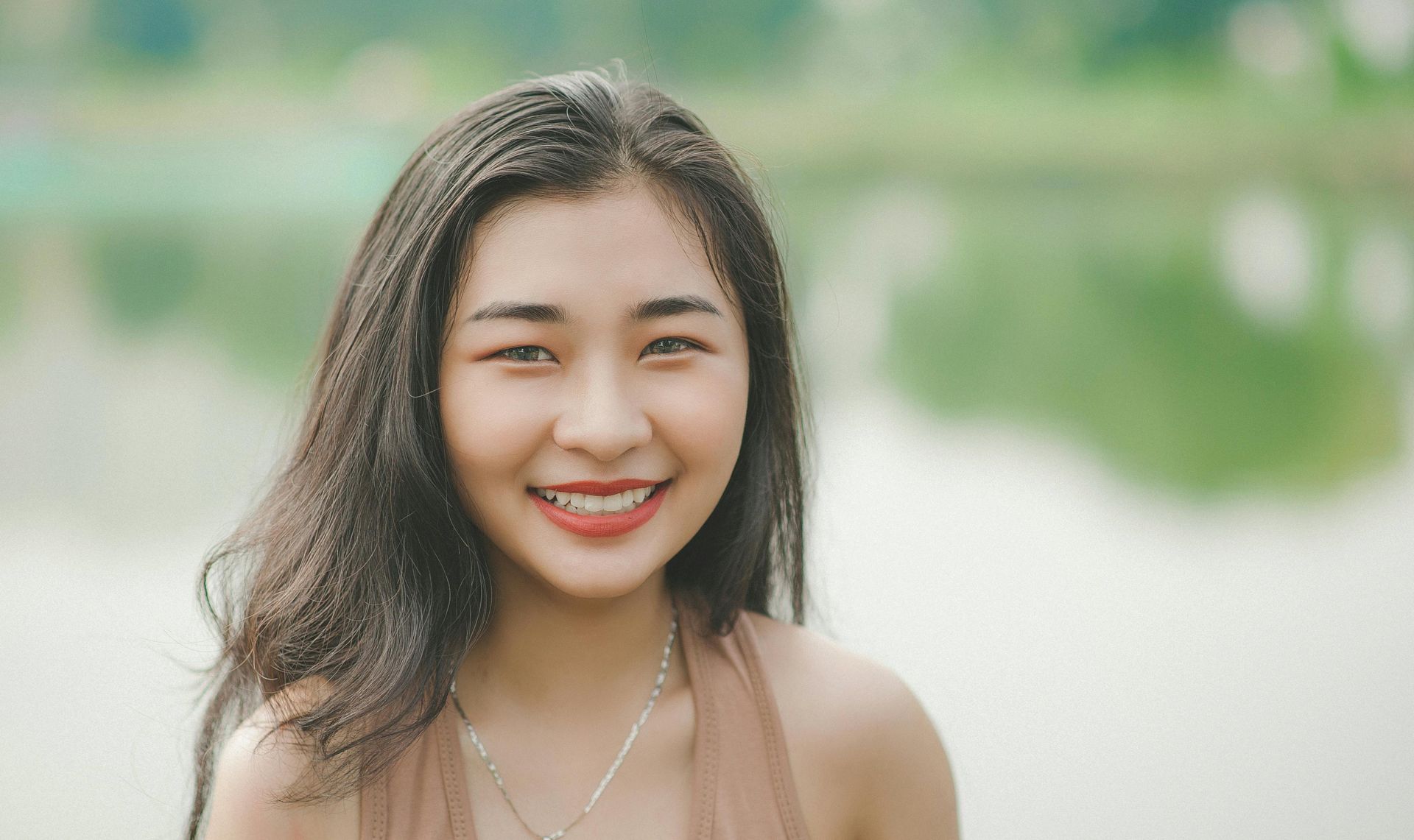 Smiling Asian woman with long dark hair outdoors, near a body of water, wearing a light brown halter top.