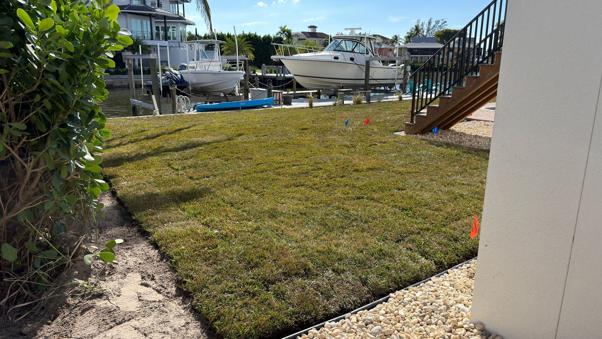 A rectangular patch of mowed grass beside a house, with a canal, boats, and stairs in the background.