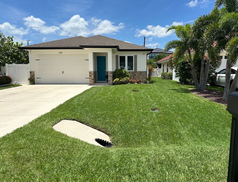 A single-story house with a white garage door, teal front door, and green lawn on a sunny day.