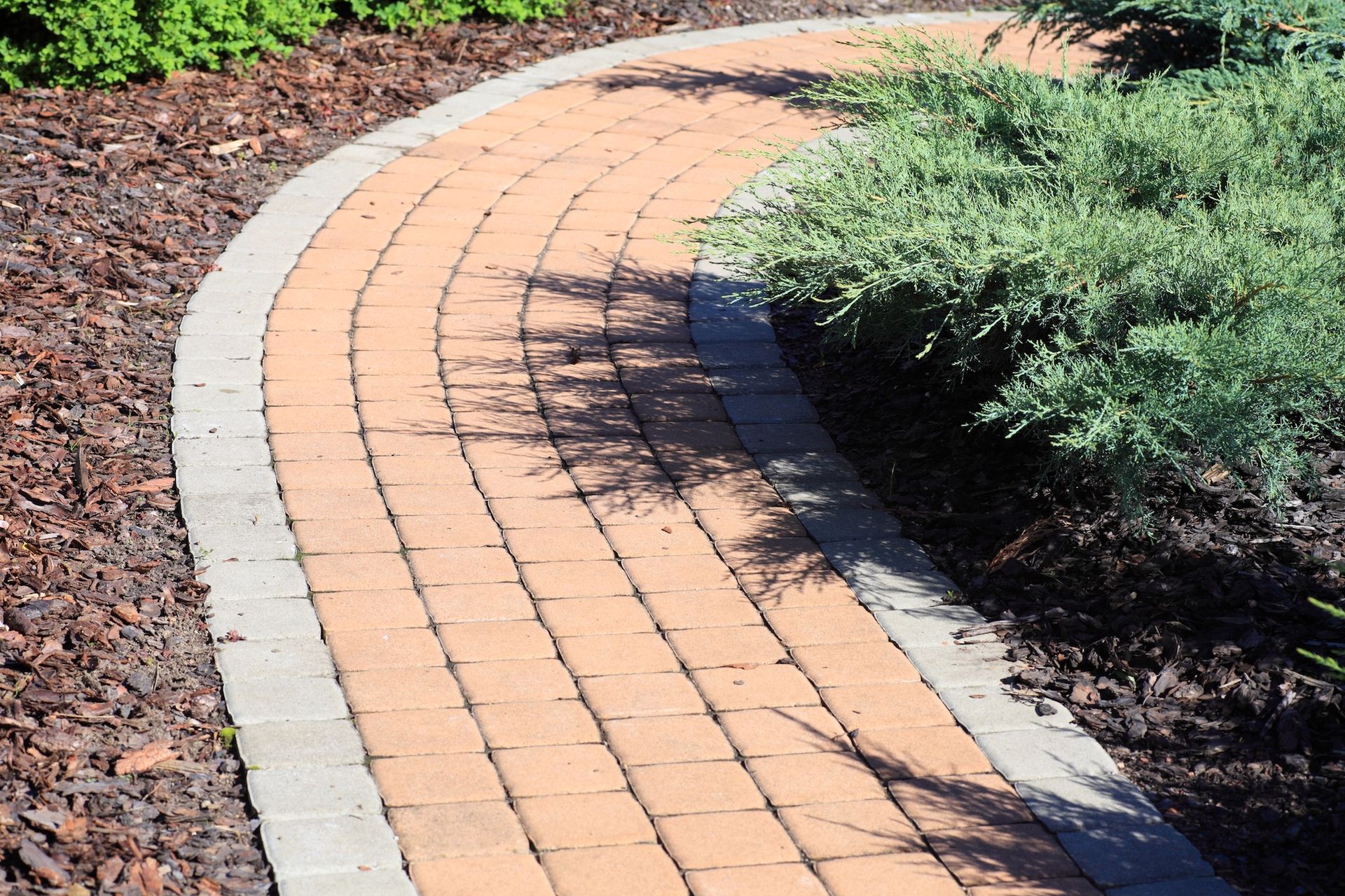 Brick pathway curves through a garden, bordered by darker edging and lush green shrubs.