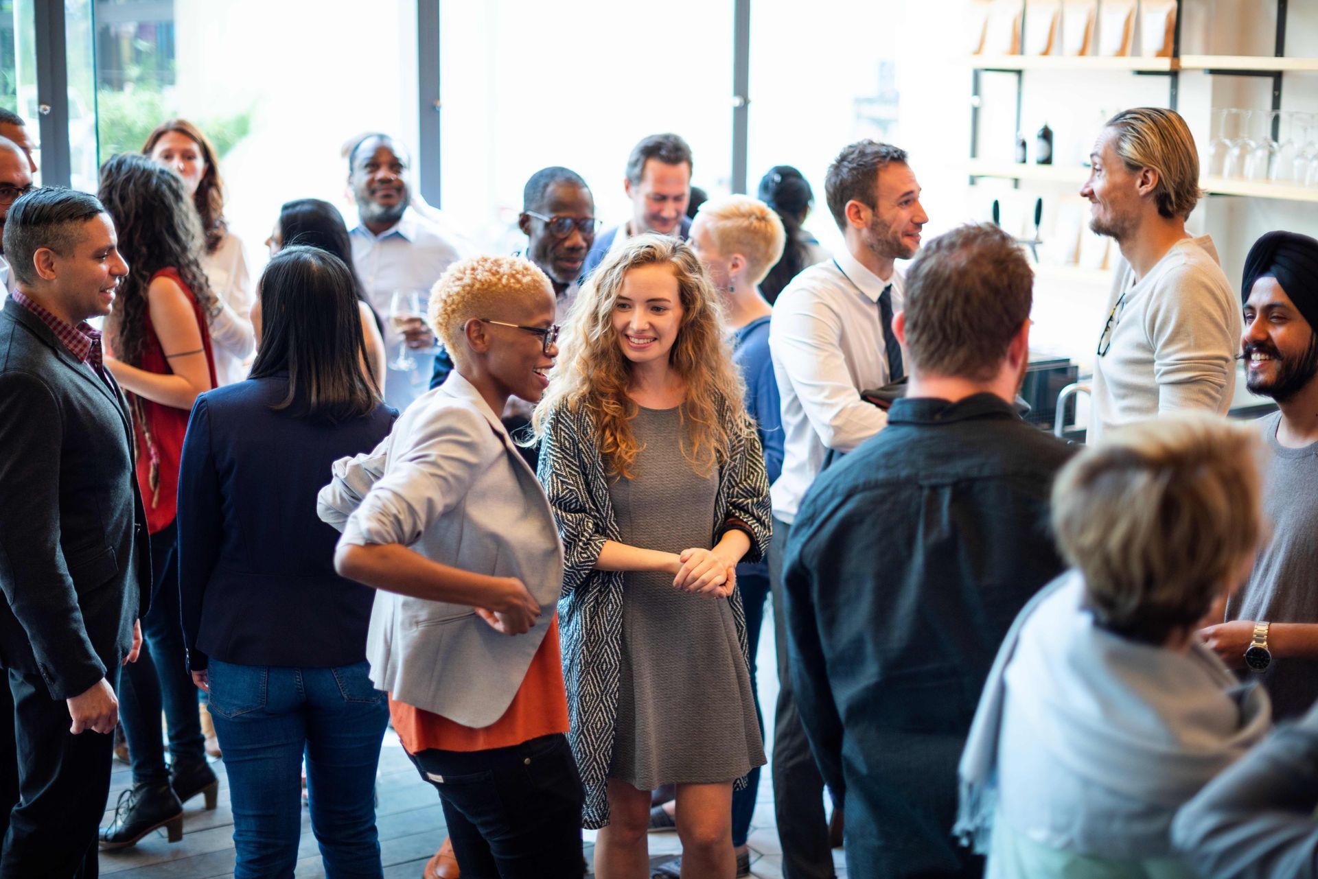 People socializing at an event, with diverse individuals in various attire, some holding drinks, in a modern, well-lit space.