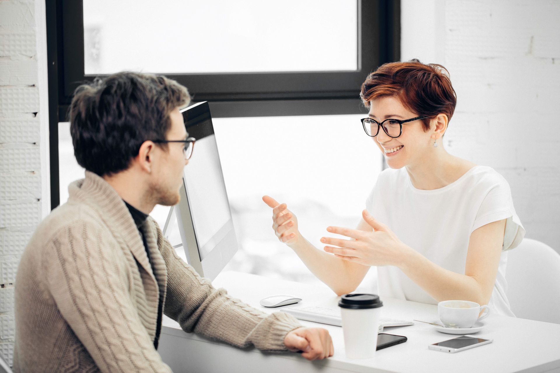 Two people sit at a desk, conversing. Woman smiles, gesturing. Man in glasses listens. White room with a window.