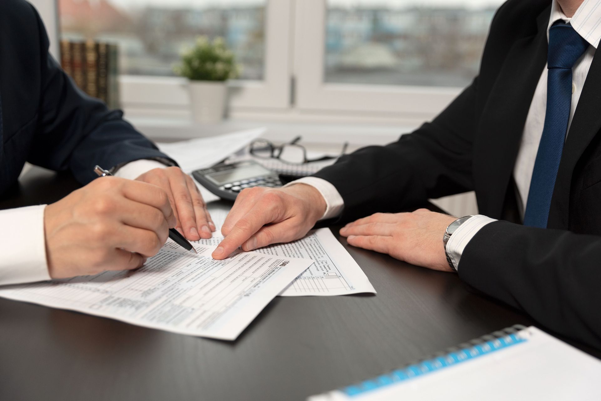 Two businesspeople in suits reviewing paperwork at a desk. One points to a document while the other holds a pen.